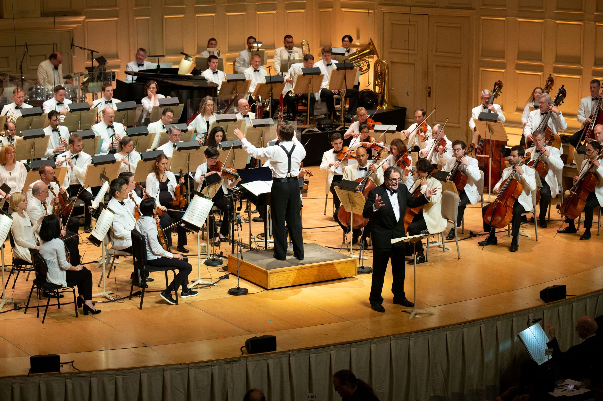An orchestra performing onstage with a conductor and a narrator standing in front of them during a performance.
