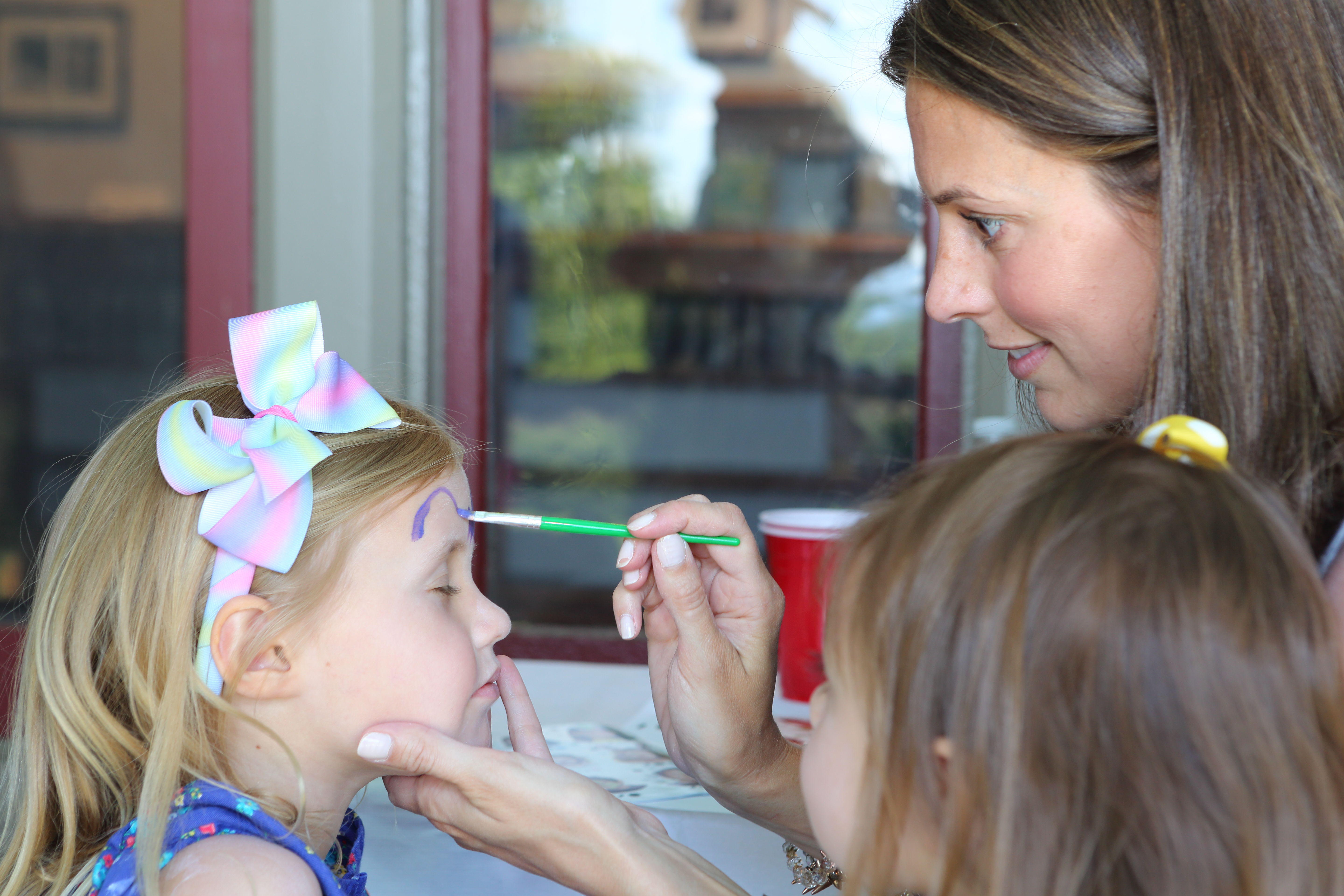 A young girl with a bright bow in her hair gets her face painted by a woman with brown hair