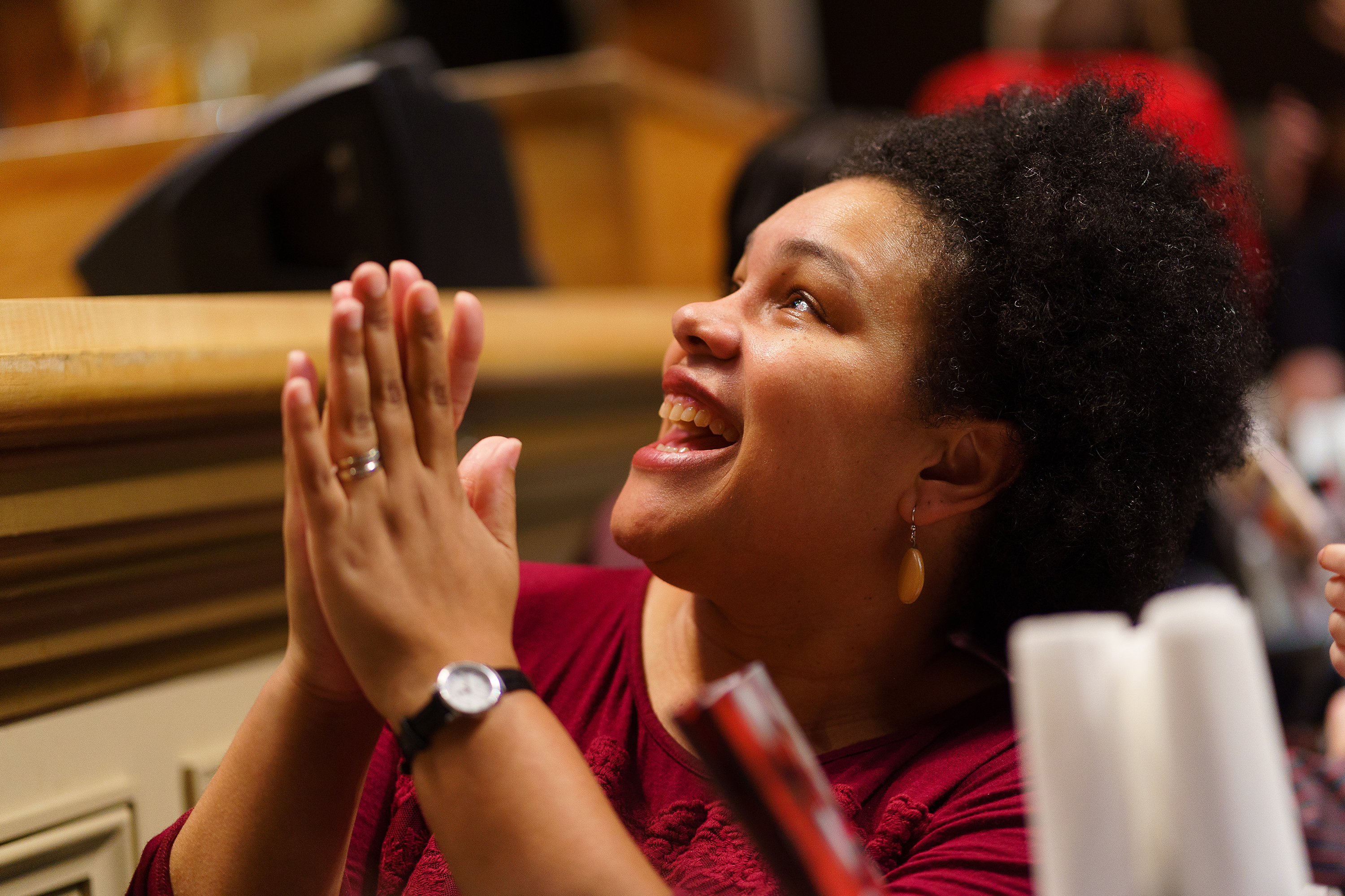 A woman in a red shirt with black, curly hair smiles and claps while sitting in Symphony Hall