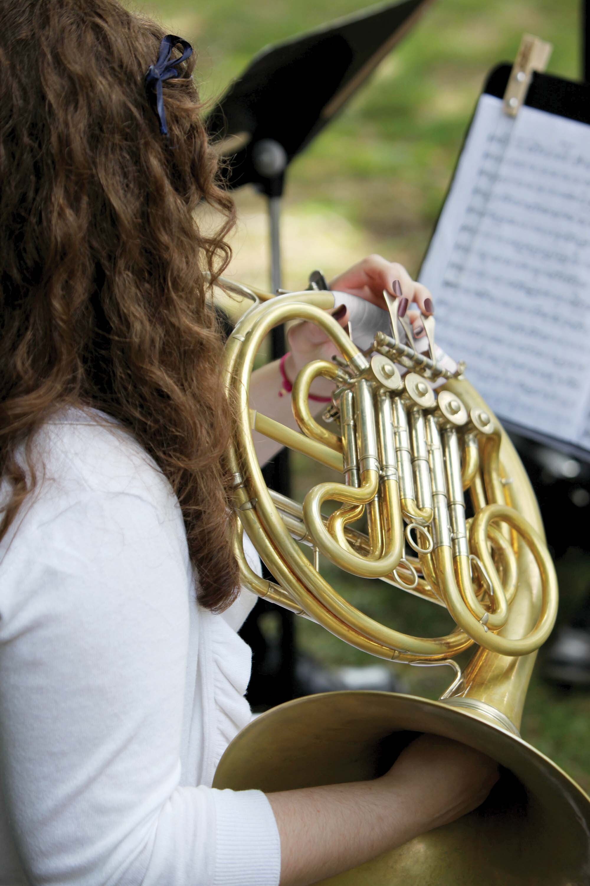 A person with long, brown curly hair plays the French horn outside