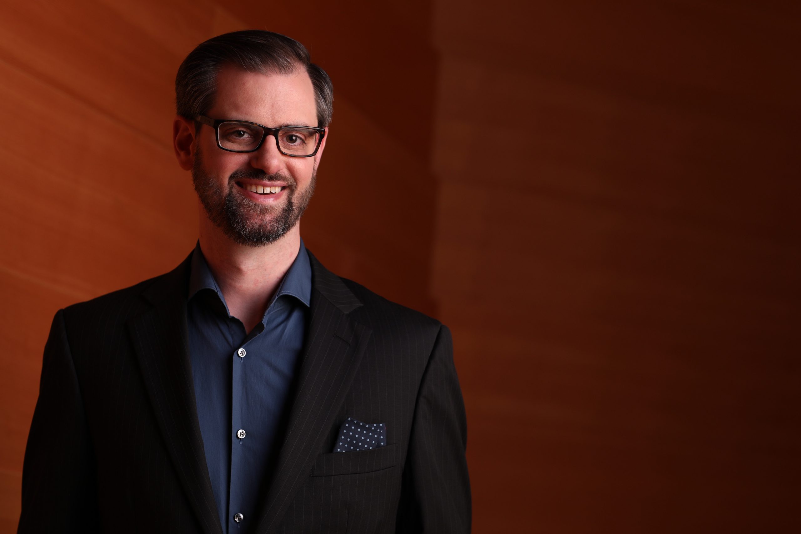 Jason Seber standing in front of a brown wall smiling and wearing a blue shirt and dark suit.