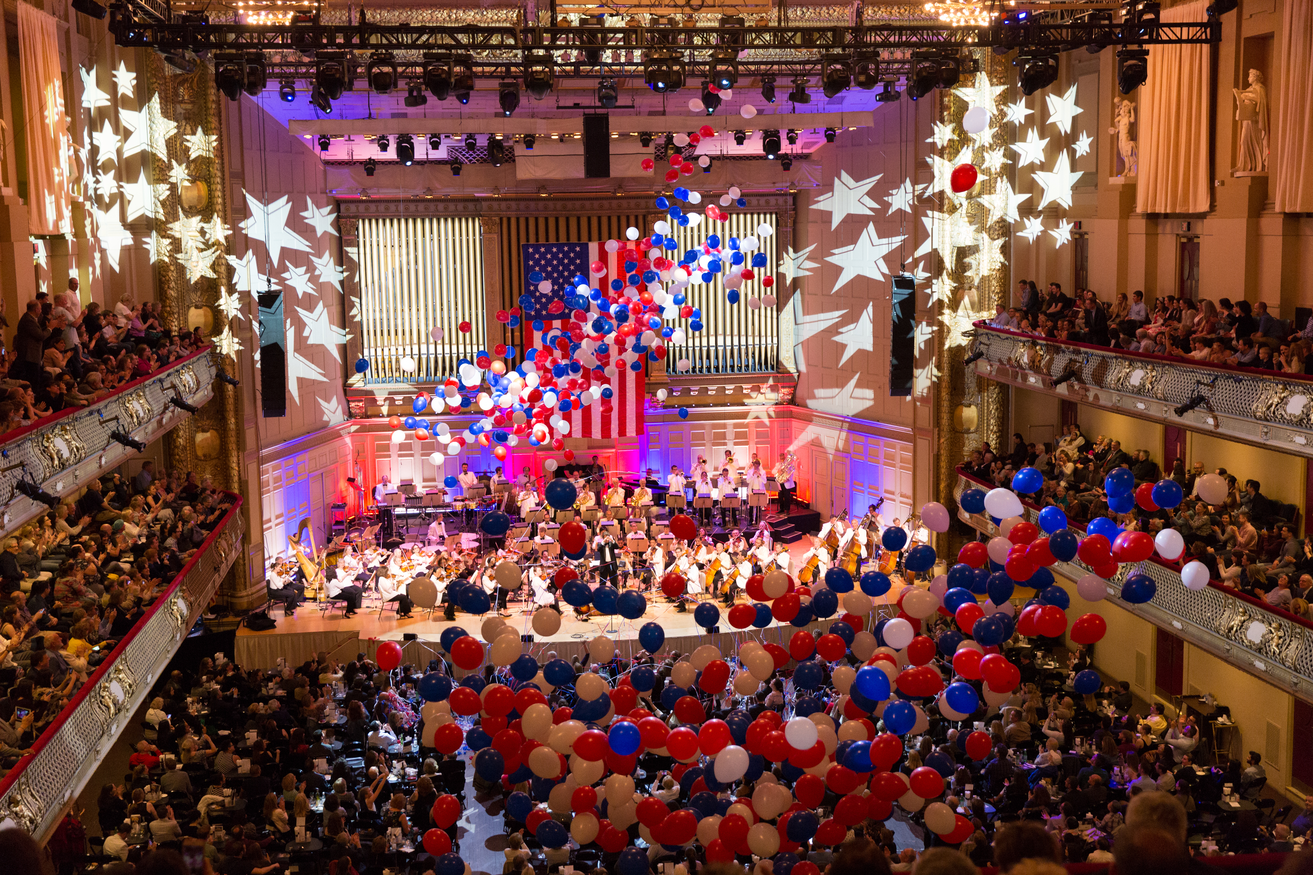 The Boston Pops perform for a full house in Symphony Hall with stars projected onstage and many red, white, and blue balloons.