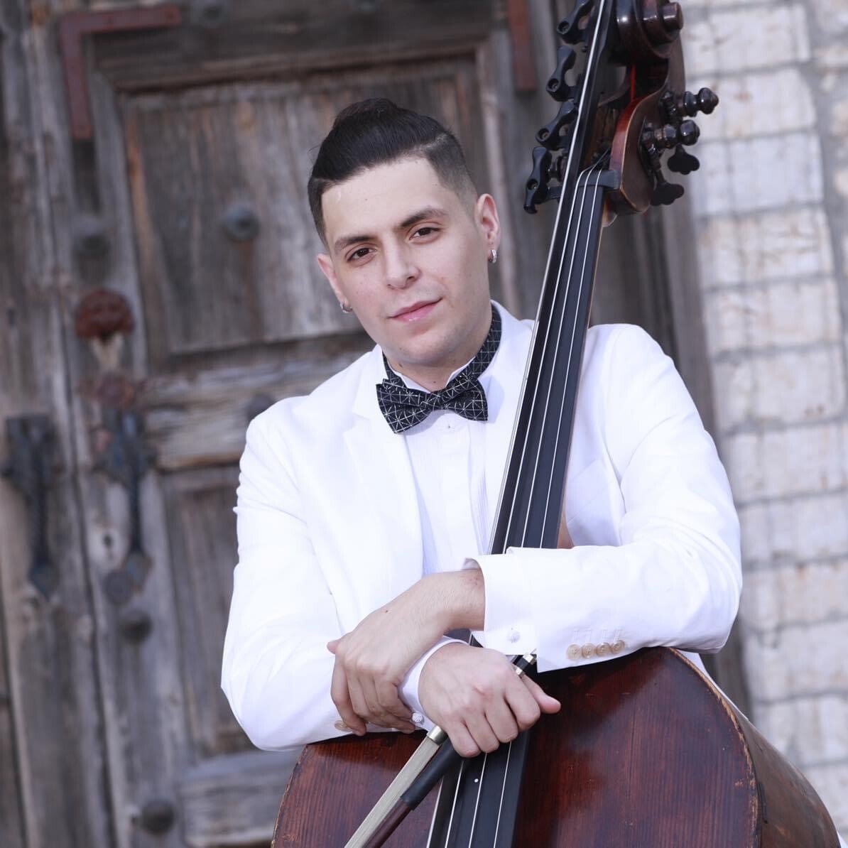 A young man in a white tuxedo with a black bowtie leans on his double bass as he gazes into the camera