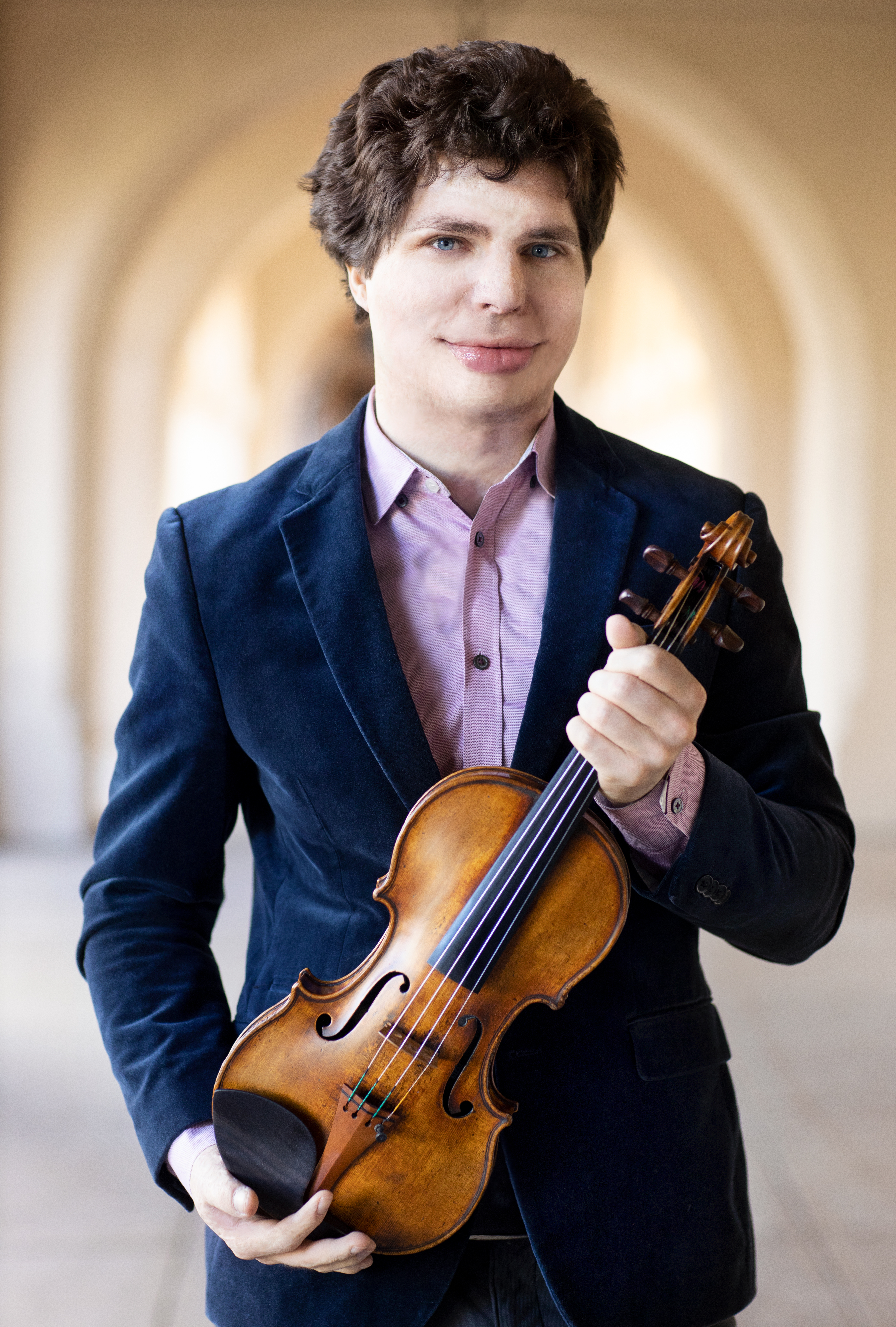 Augustin Hadelich wearing a purple shirt and blue suit jacket, holding a violin close with both hands.