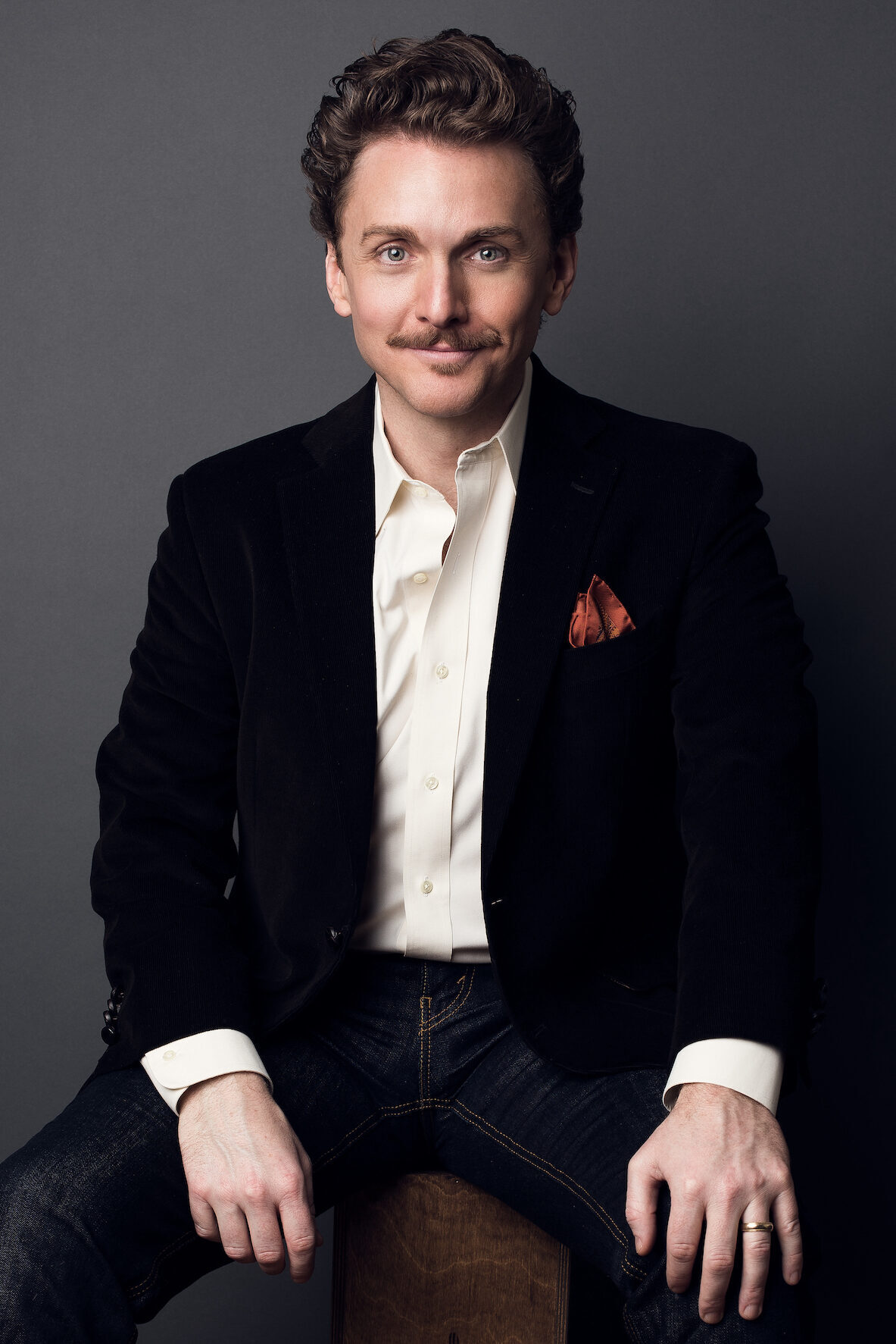 Jason Danieley seated on a box with a dark blue suit, white shirt, and red pocket square