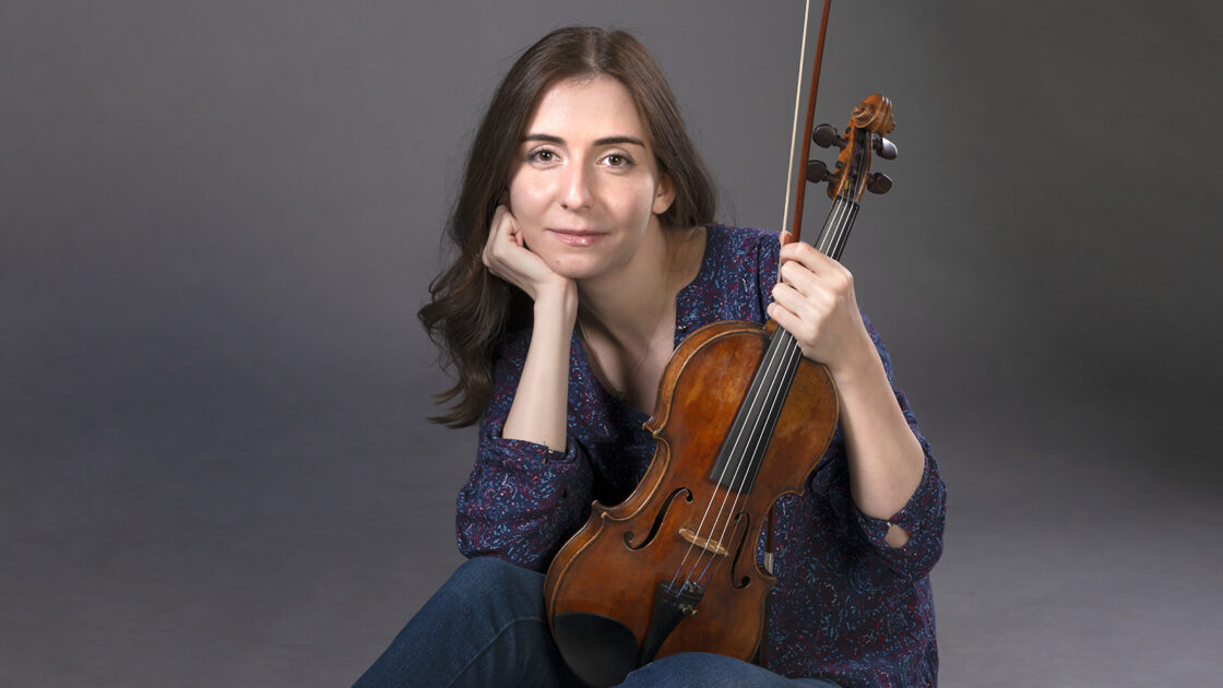 Diana Adamyan sitting down in a blue gown holding her violin and bow.