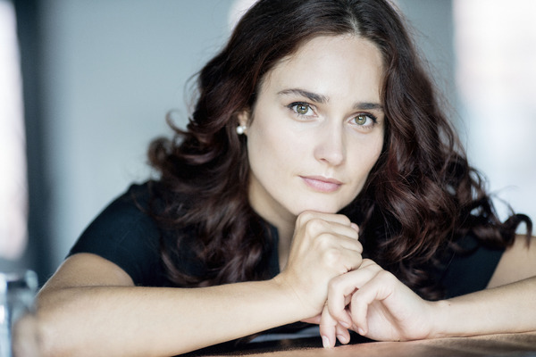 A headshot of Anna Vinnitskaya leaning on a piano with arms crossed