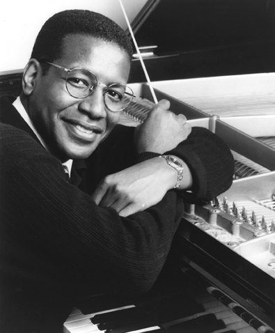 Black and white photo of Charles Floyd sitting at a piano, holding a conductor's baton.