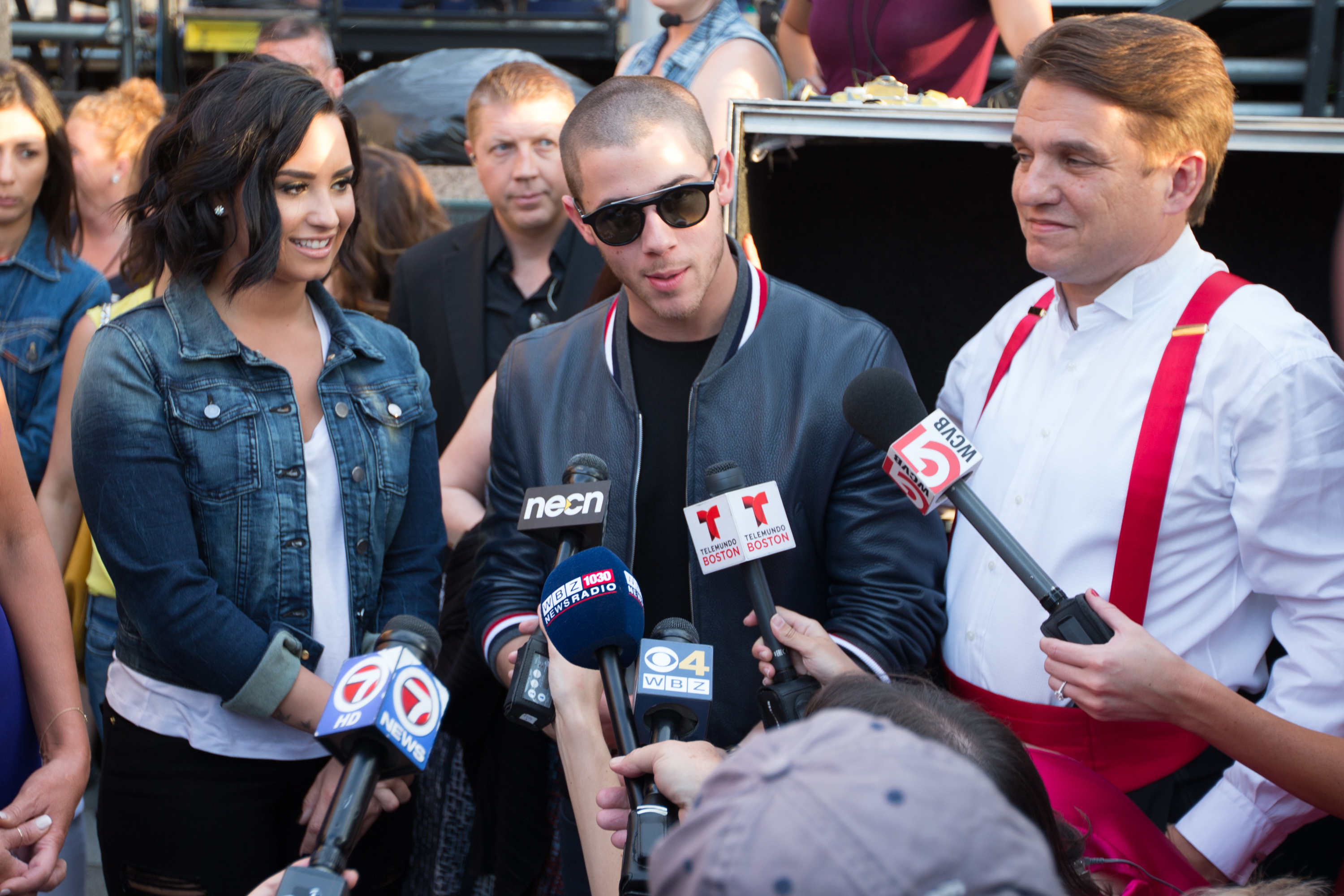 Demi Lovato, Nick Jonas, and Keith Lockhart stand close together with a crowd gathered behind them as Jonas speaks into microphones held by several reporters
