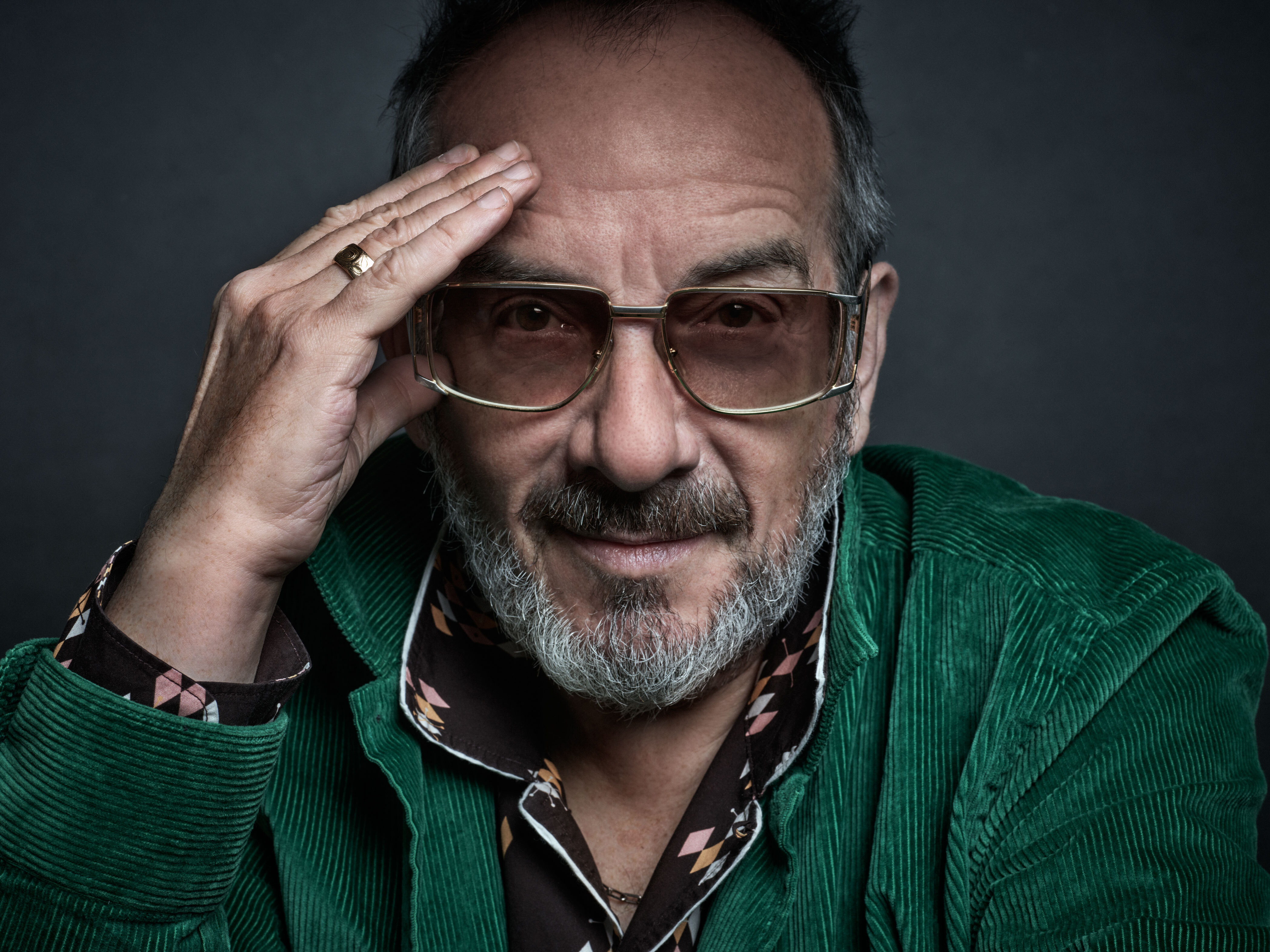 A closeup headshot of Elvis Costello with his hand on his temple, and wearing a green corduroy jacket and a black open collar shirt