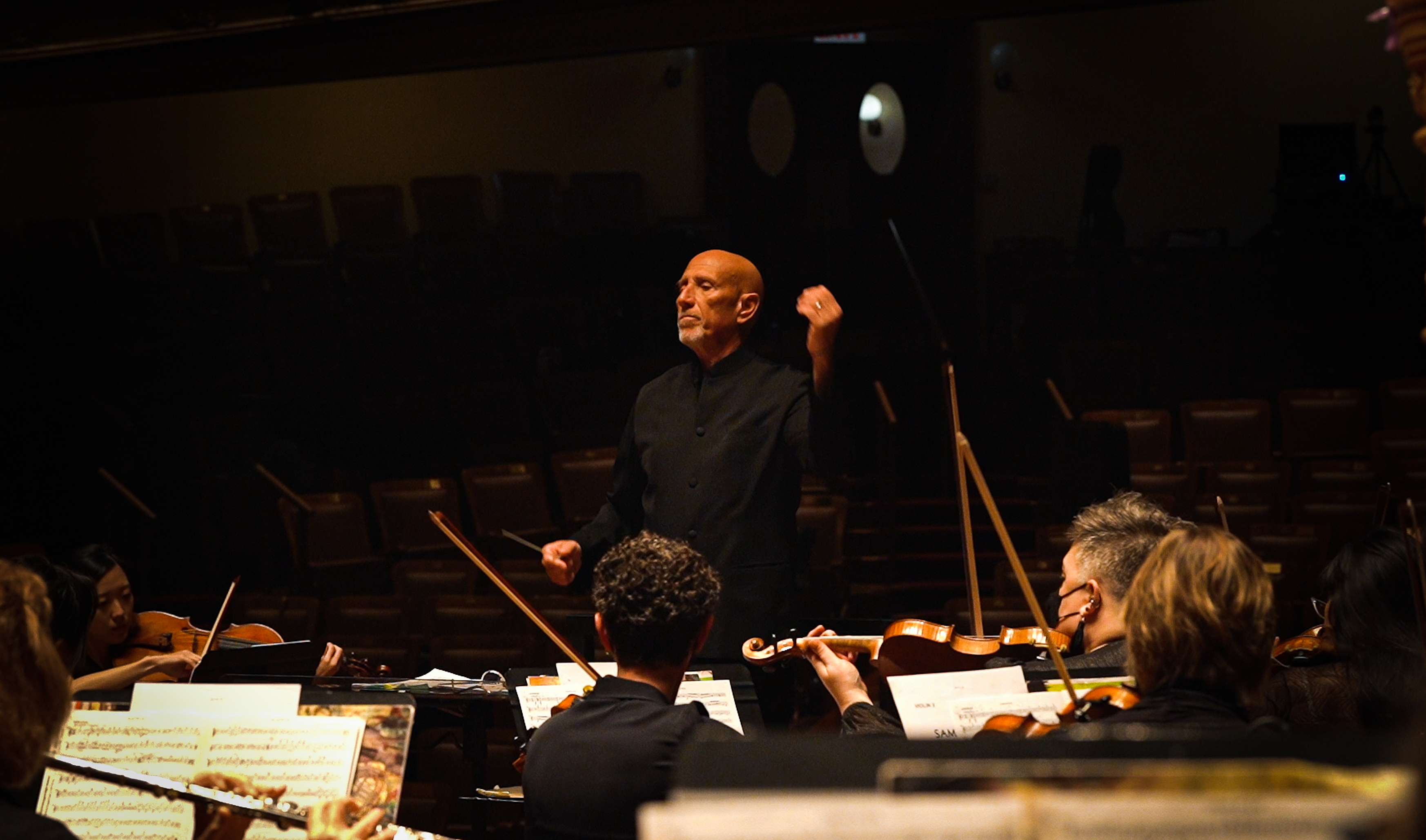 Francisco Noya wearing black, standing on the conductor podium, conducting an orchestra while holding a baton.