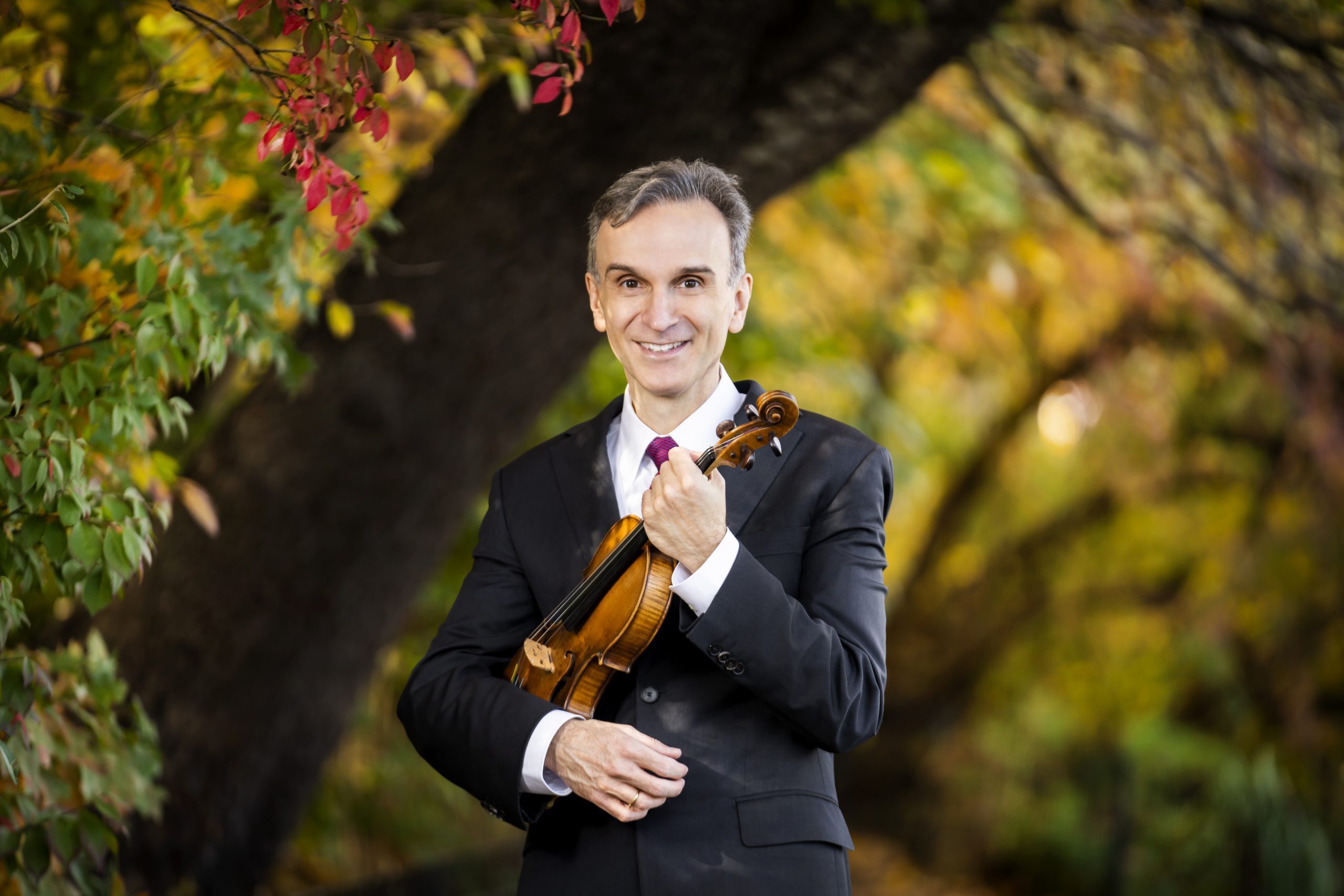 Gil Shaham wearing a dark suit holding his violin and standing in front of a tree.