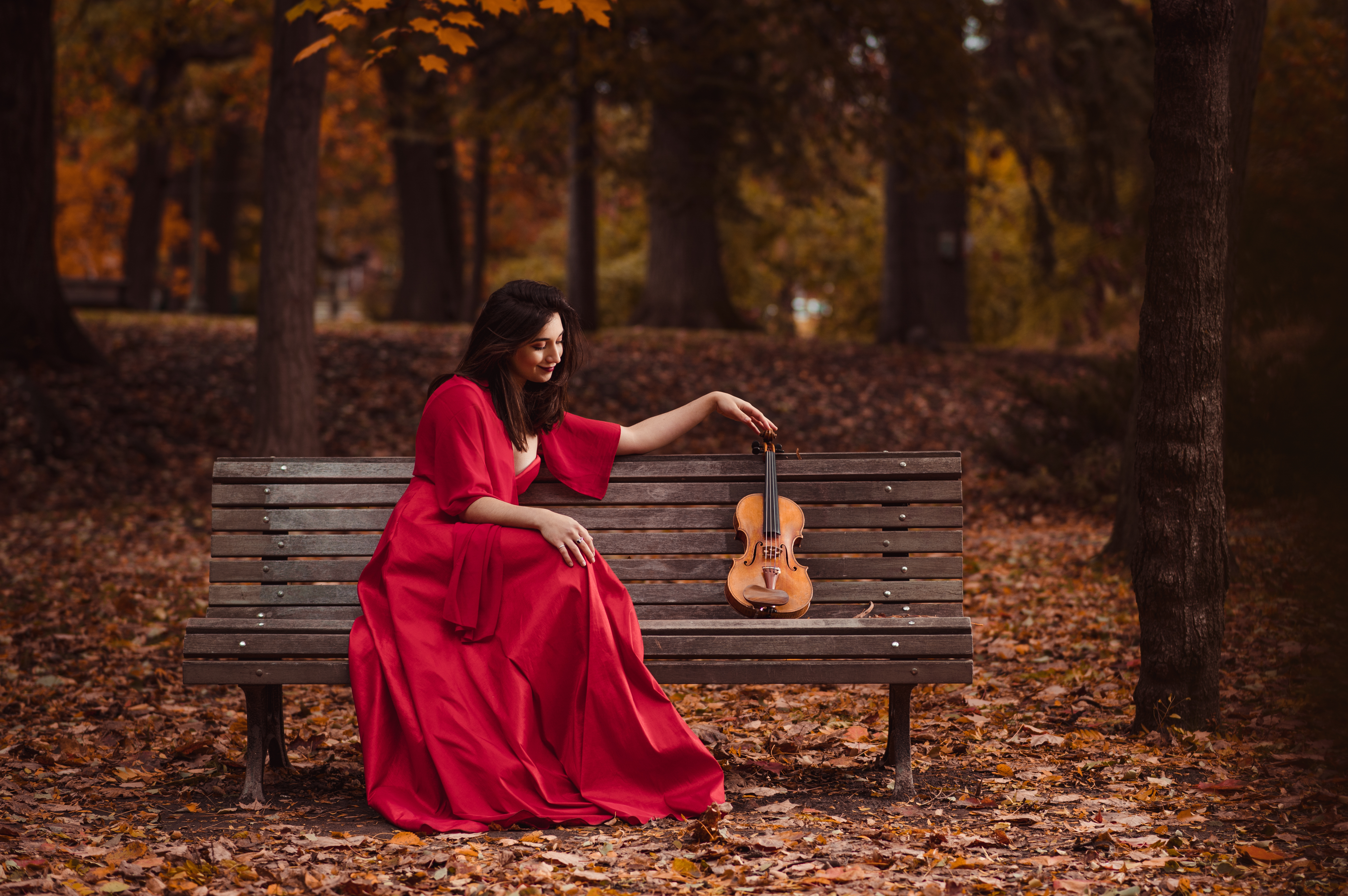 Isabella Perron sitting on a park bench wearing a long red dress, with her arm outstretched touching the neck of her violin that is next to her on the bench.
