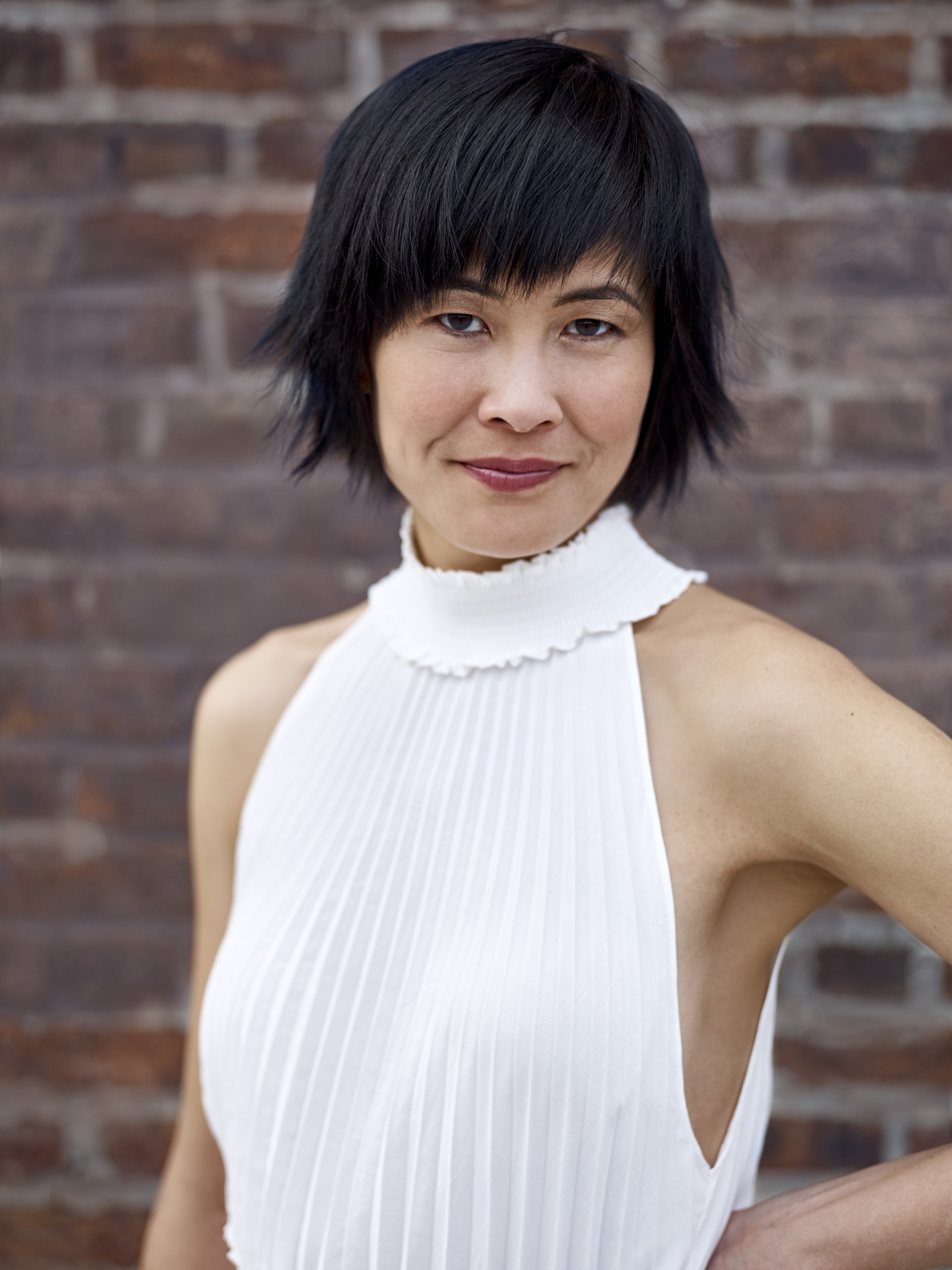 Jennufer Koh posing in front of a brick wall wearing a white top.