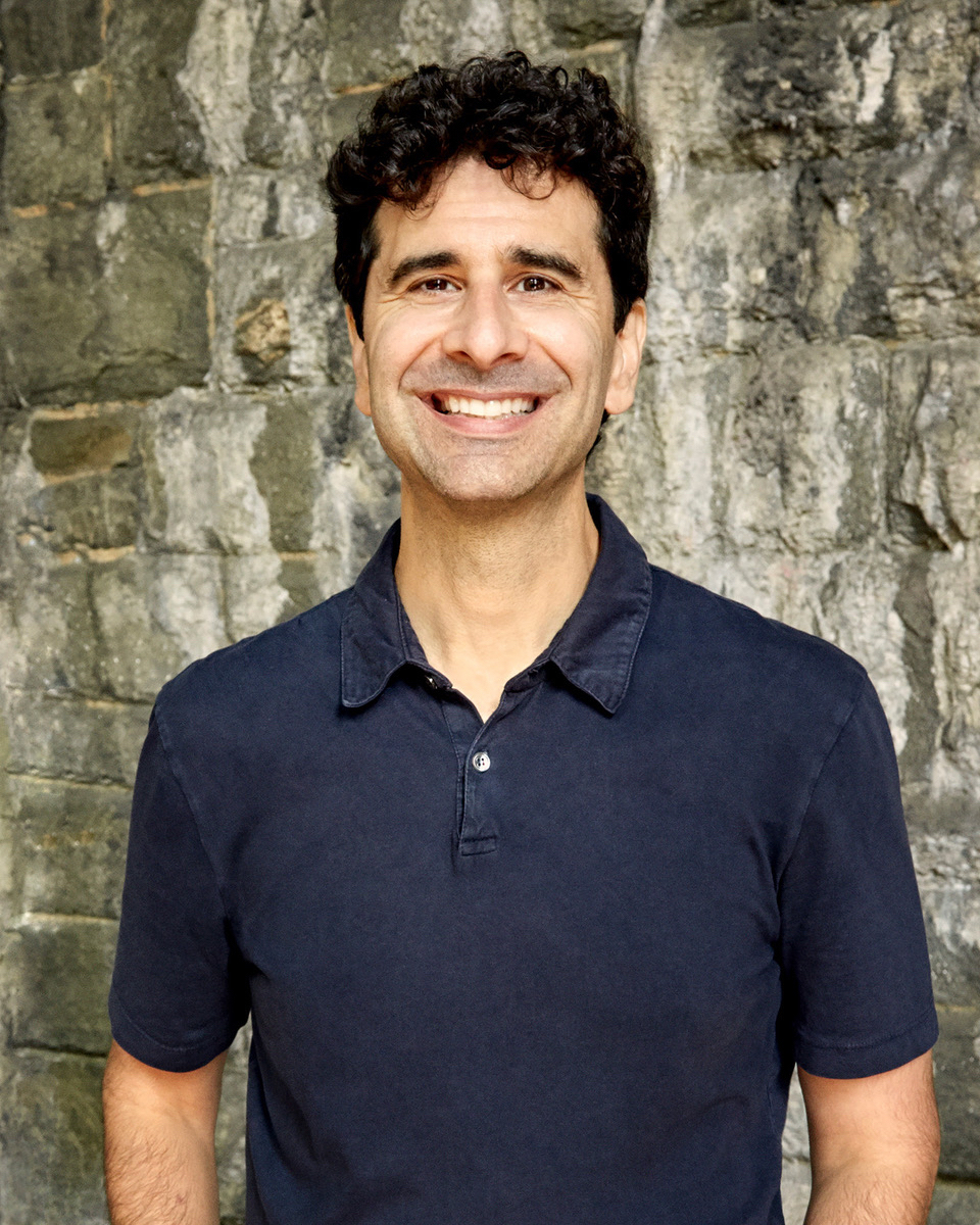 A headshot of John Carini standing against a stone wall wearing a dark blue polo
