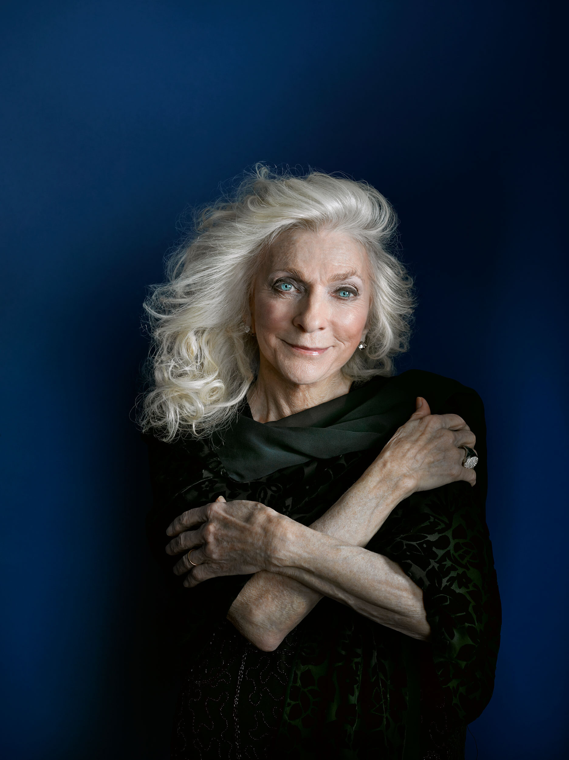 Judy Collins standing in front of a blue background with her arms crossed in front of her chest