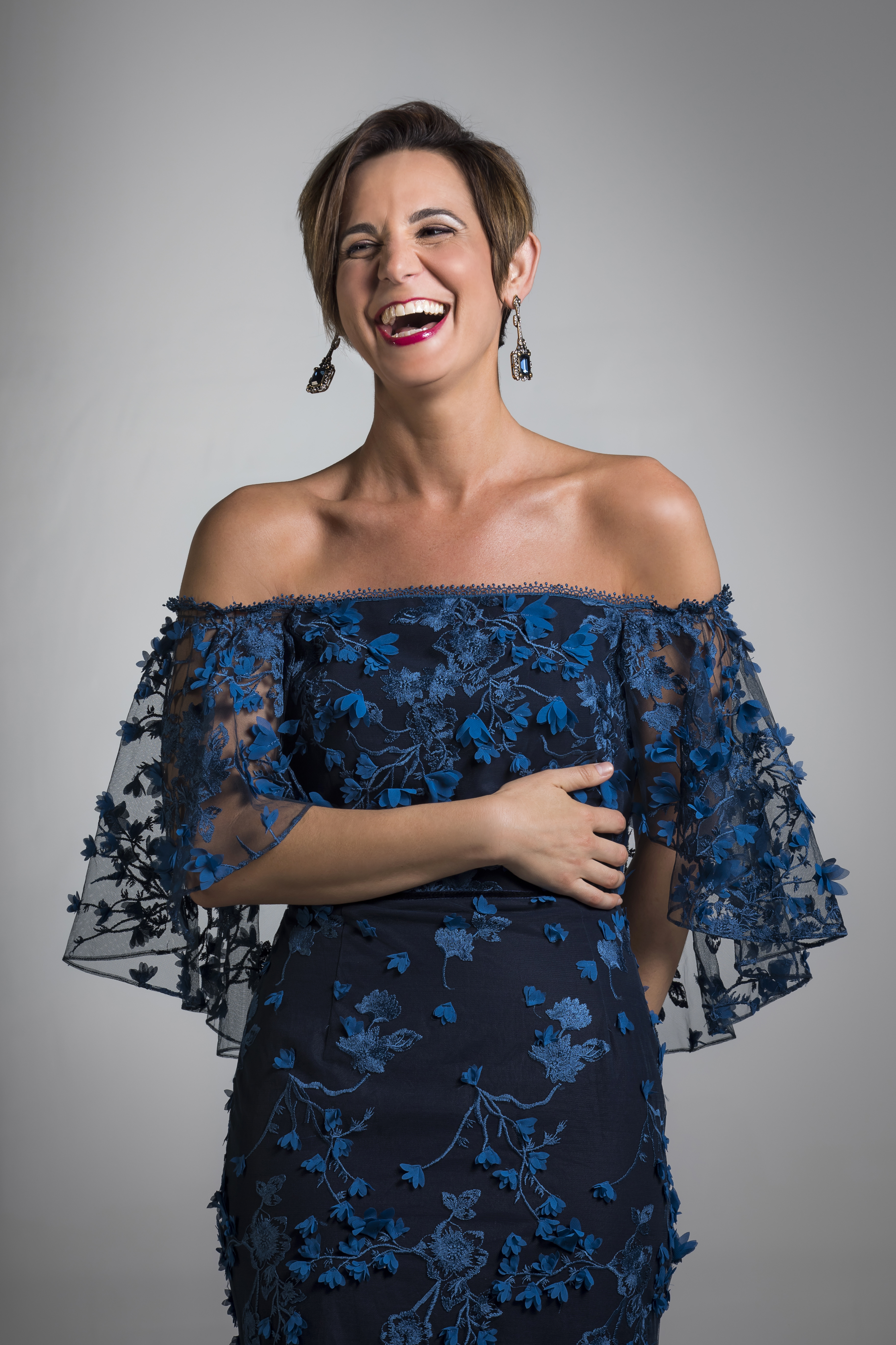 Kelley O'Connor standing in a blue dress in front of a gray background, laughing