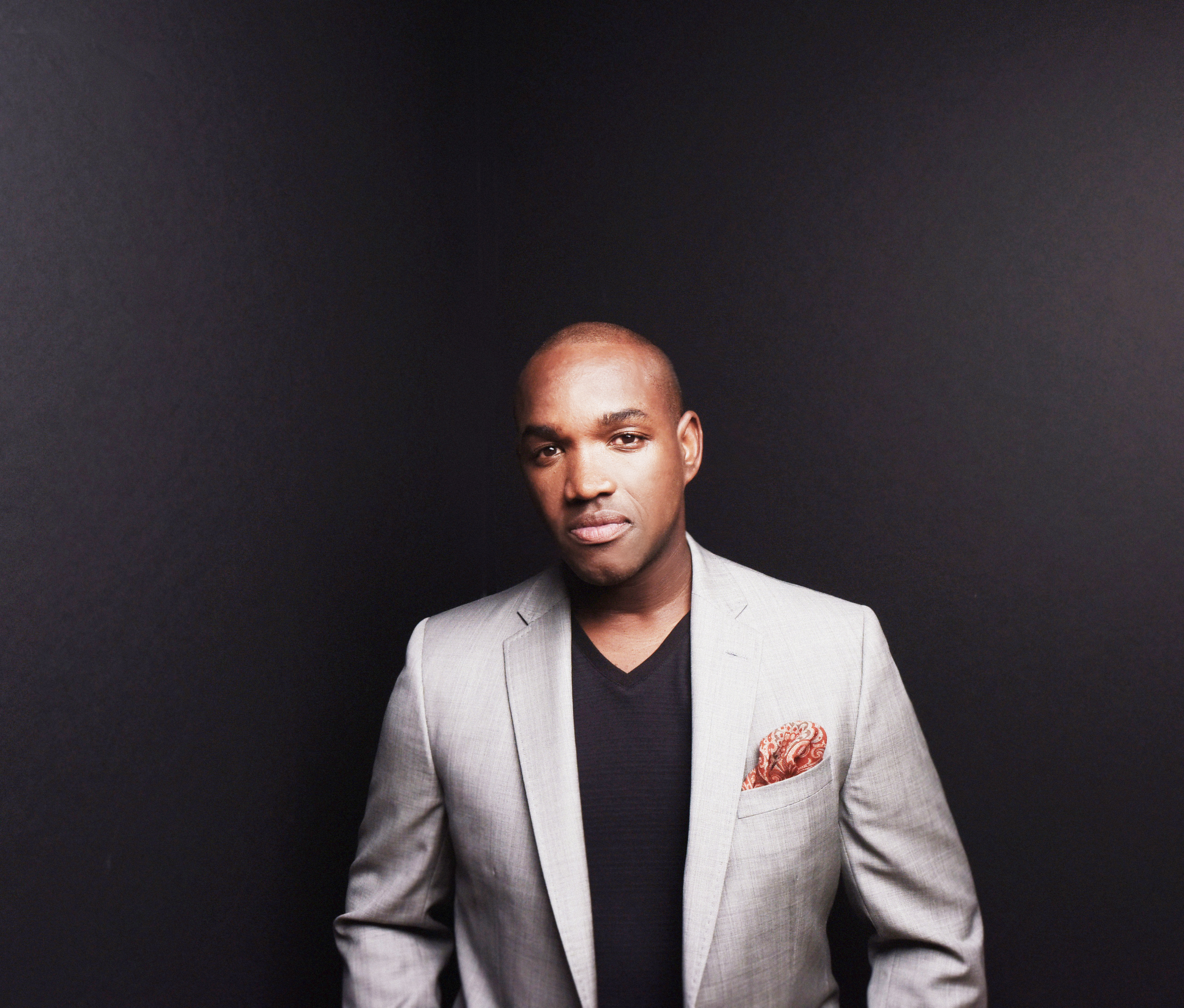 Lawrence Brownlee standing against a black background wearing a grey jacket with red pocket square