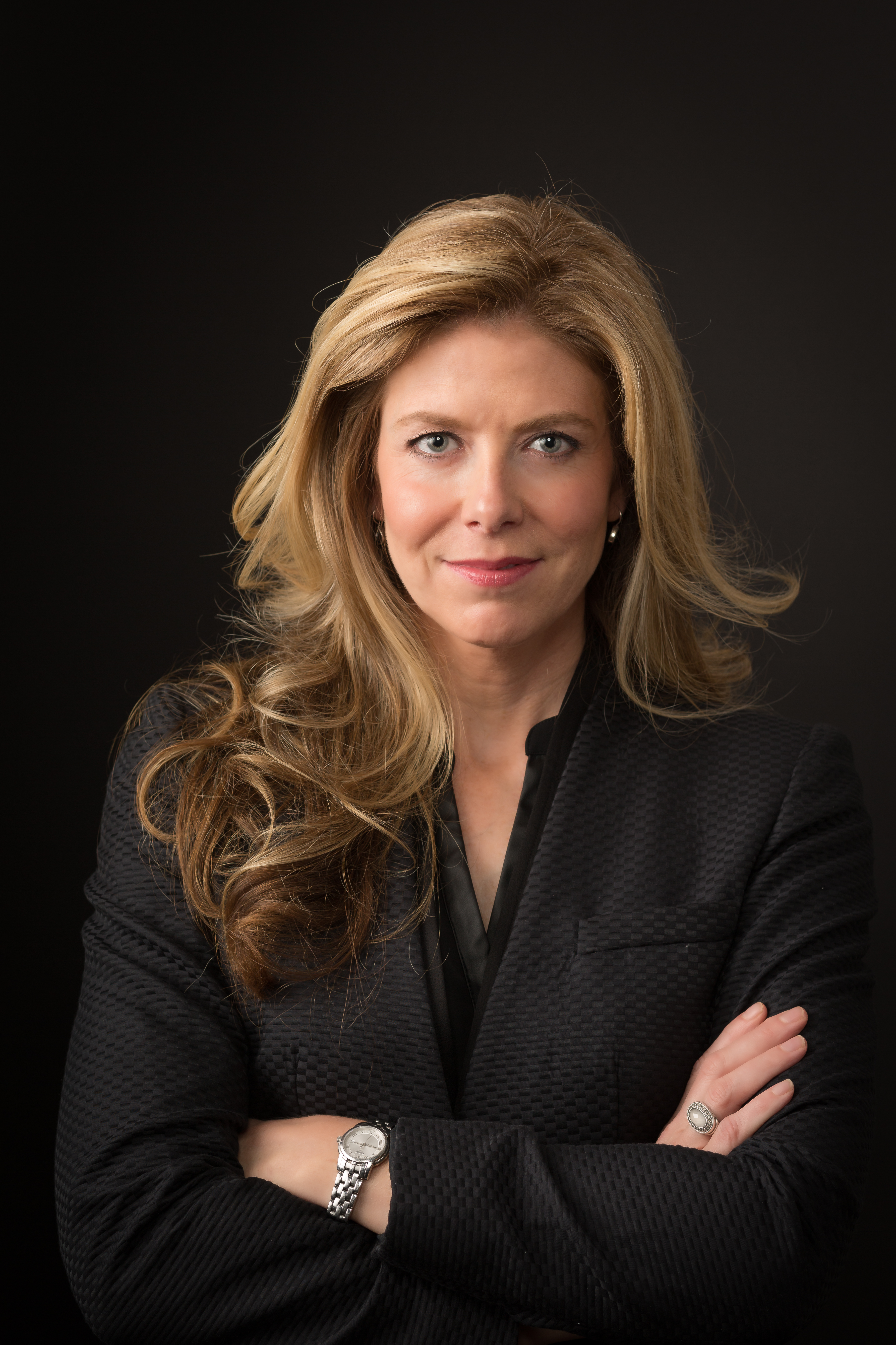 A smiling blond woman stands in front of a black background with her arms crossed in front of her