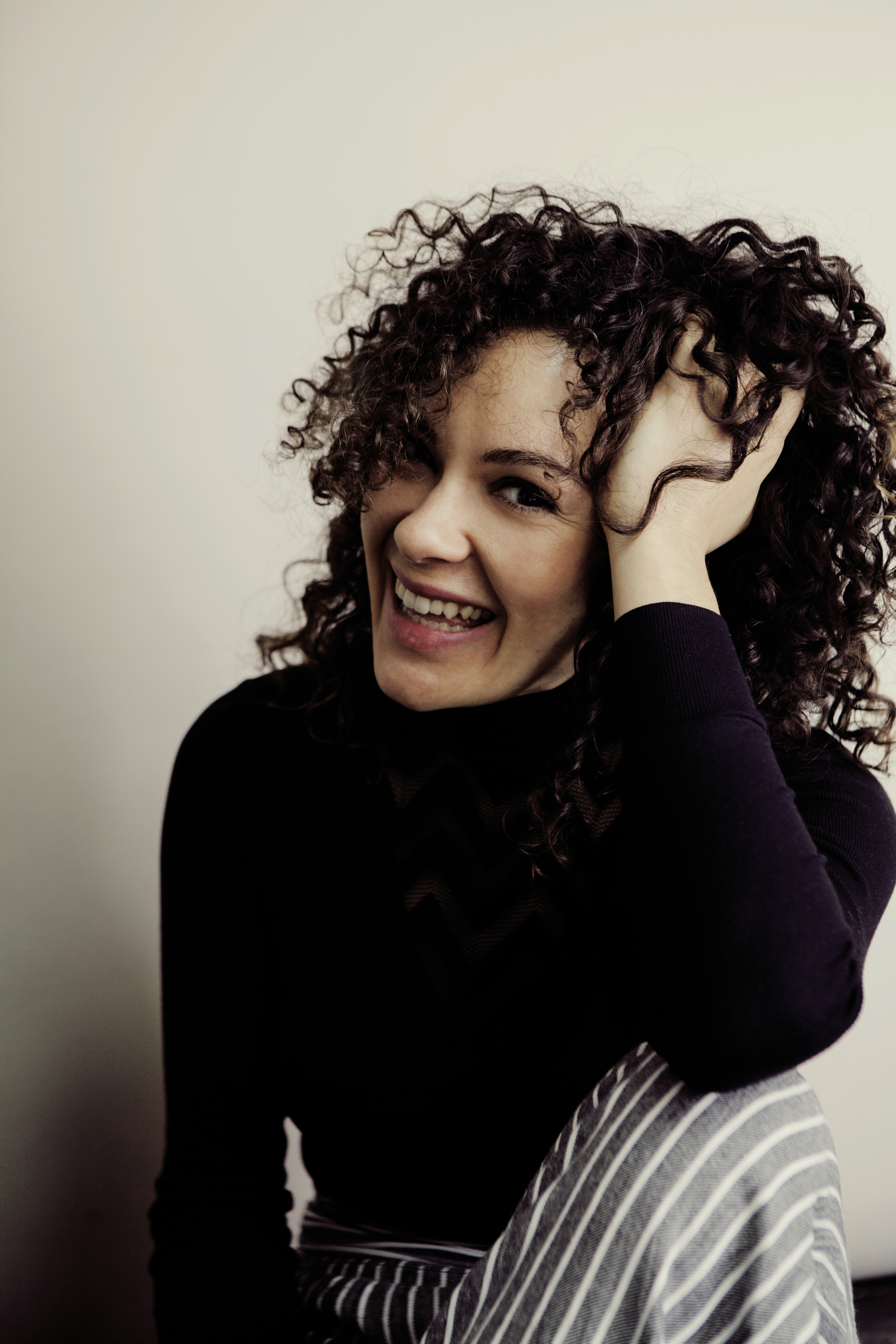 Portrait of Nora Fischer wearing a black shirt and striped pants, resting her elbow on her knee with her hand in her hair