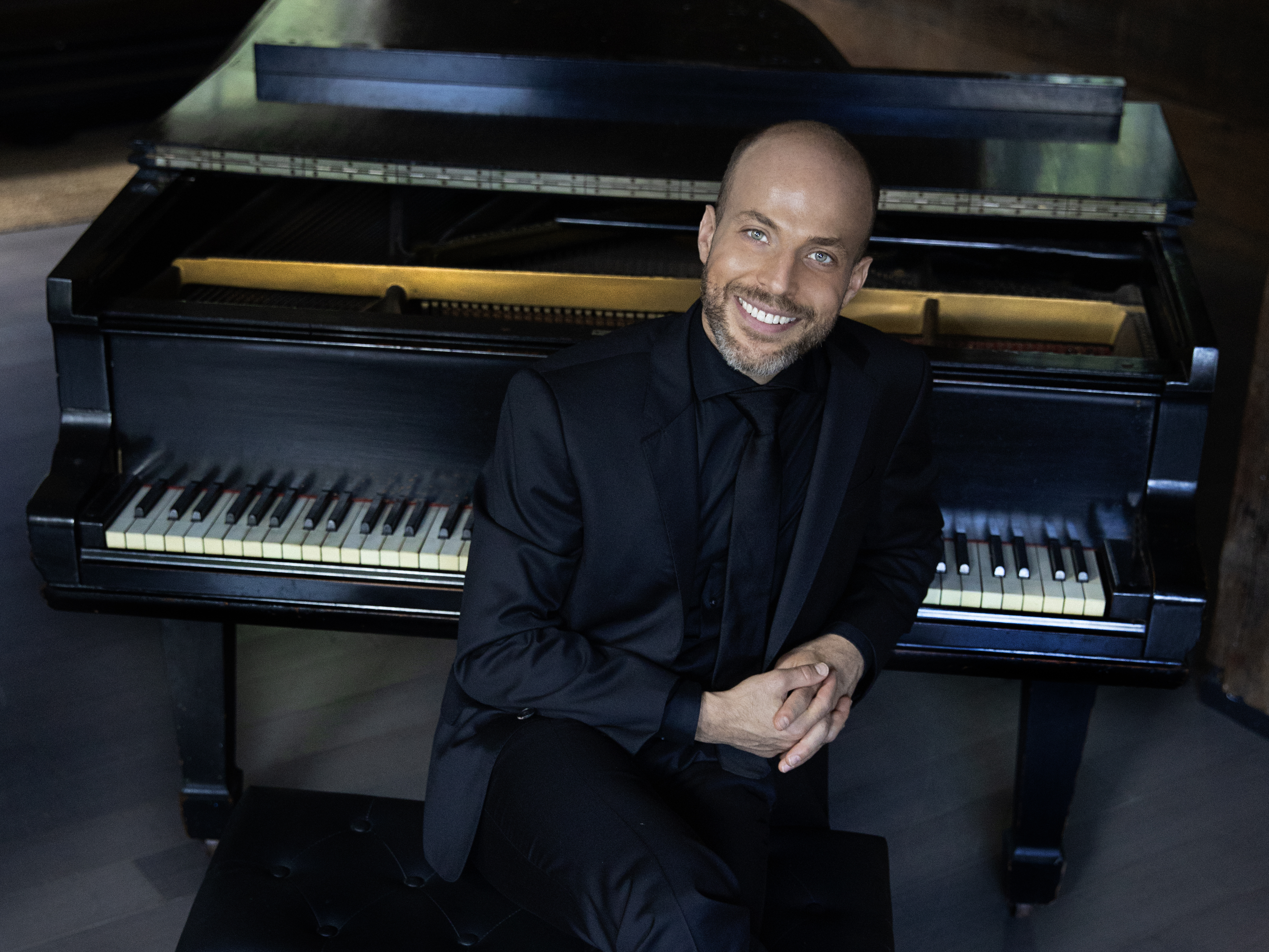 Headshot of Orion Weiss wearing a black suit and sitting in front of a piano.