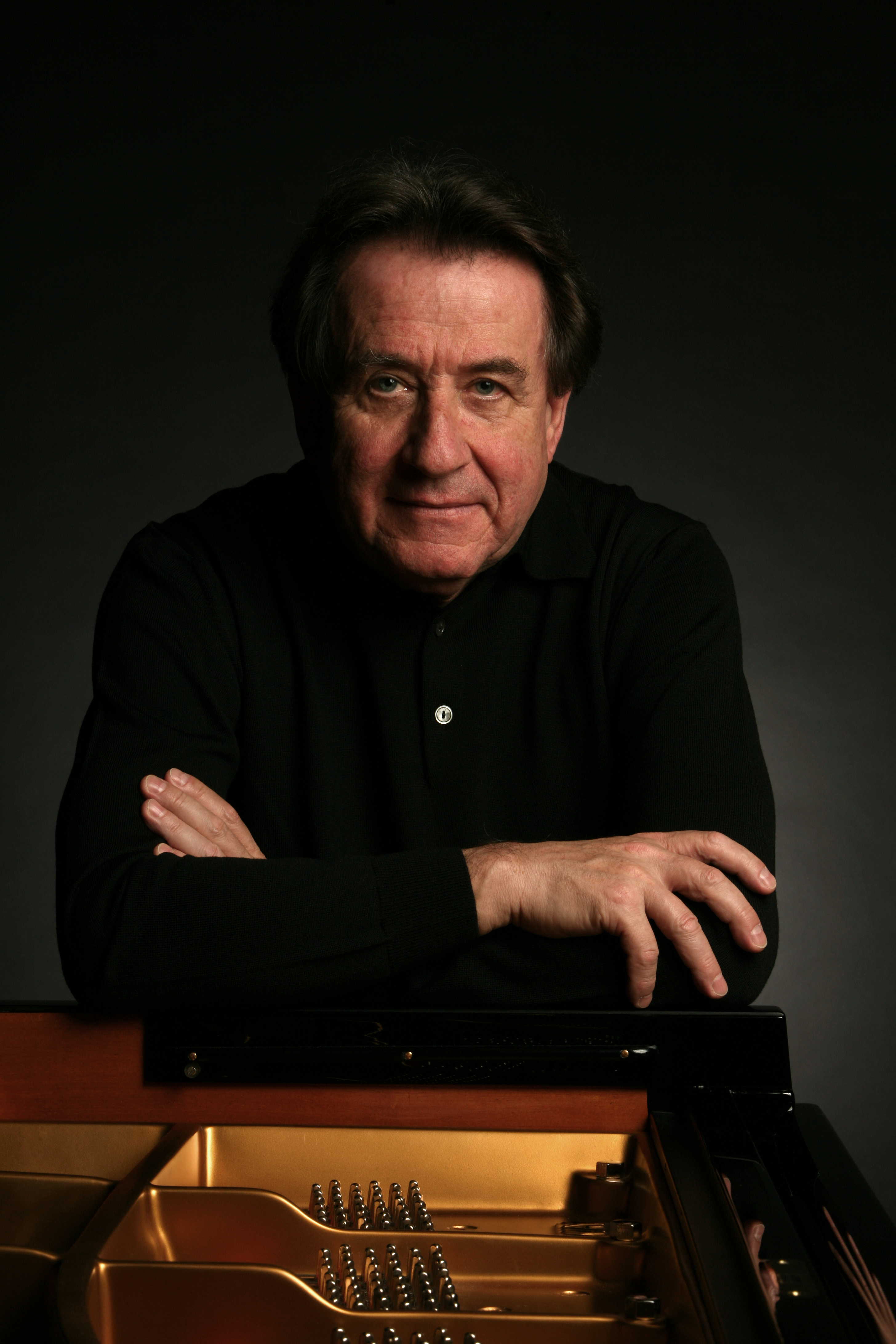 Rudolf Buchbinder leaning on the top of an open piano with arms crossed, smiling