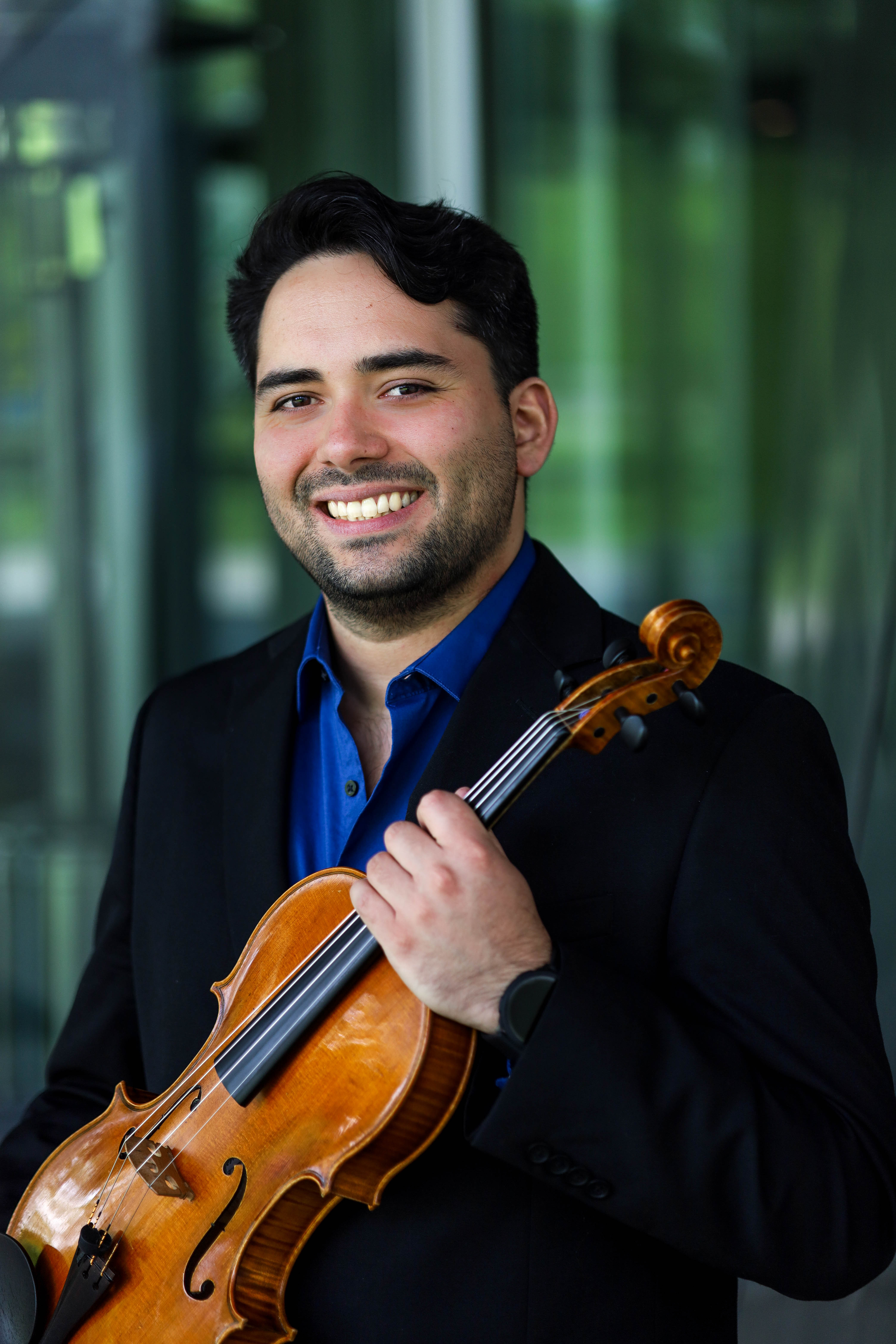 A man in a suit with short, dark hair holds his viola and smiles for a photo