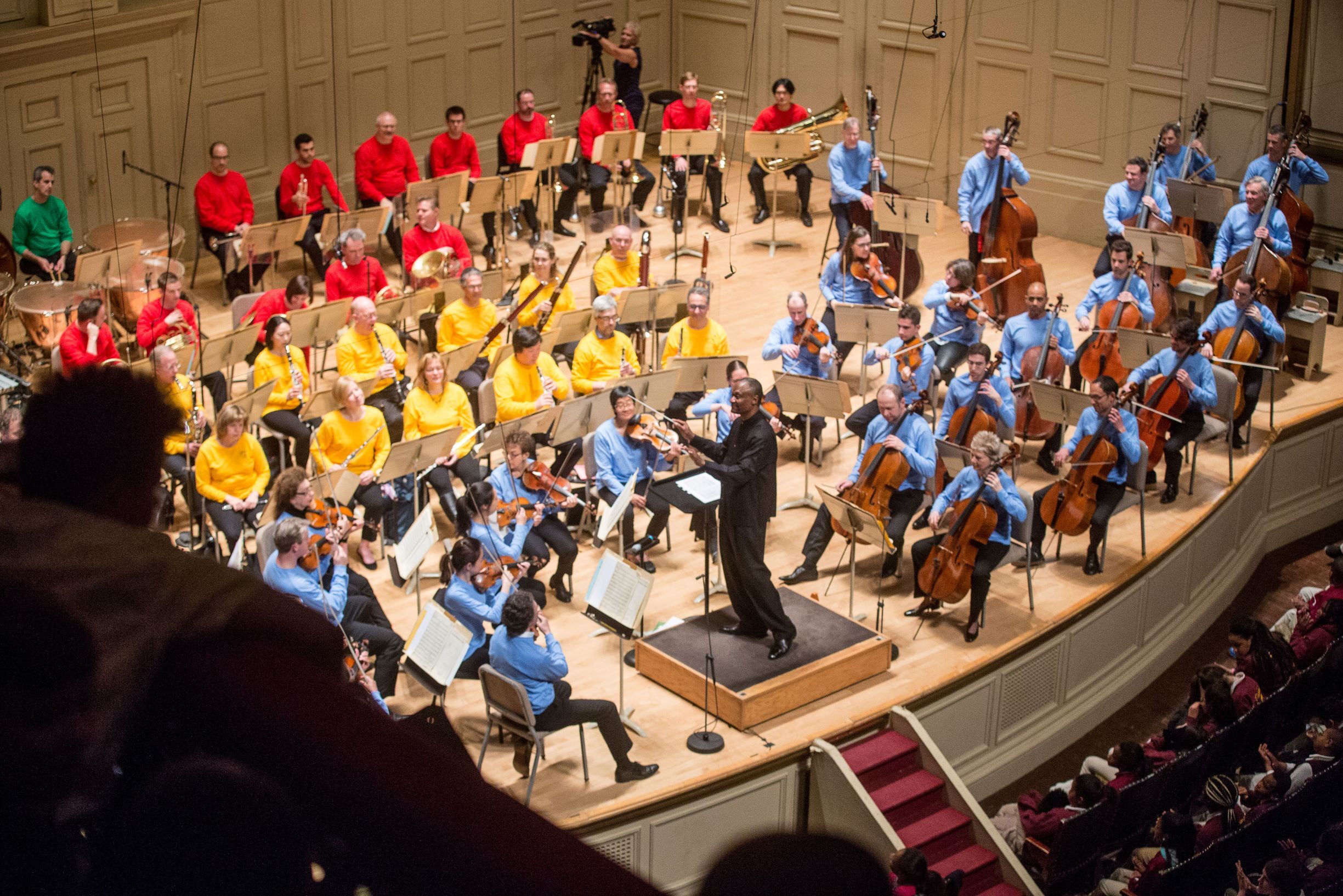 Thomas Wilson conducts BSO musicians who are all wearing colorful shirts. A student watches from the balcony.