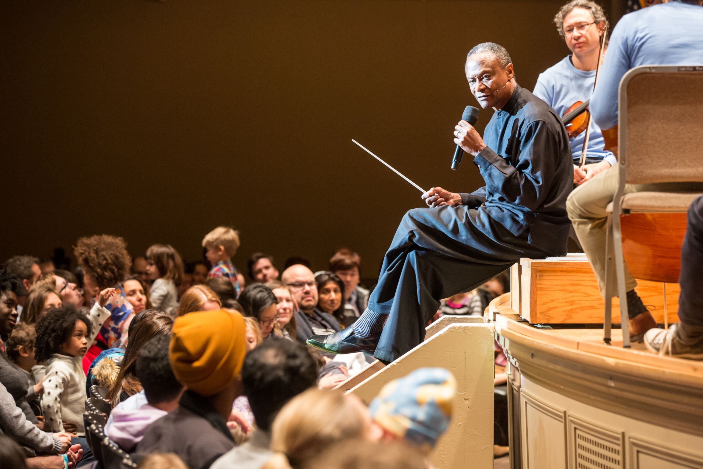 Thomas Wilkins sitting on the edge of the stage during a performance talking to an audience of children and their families.