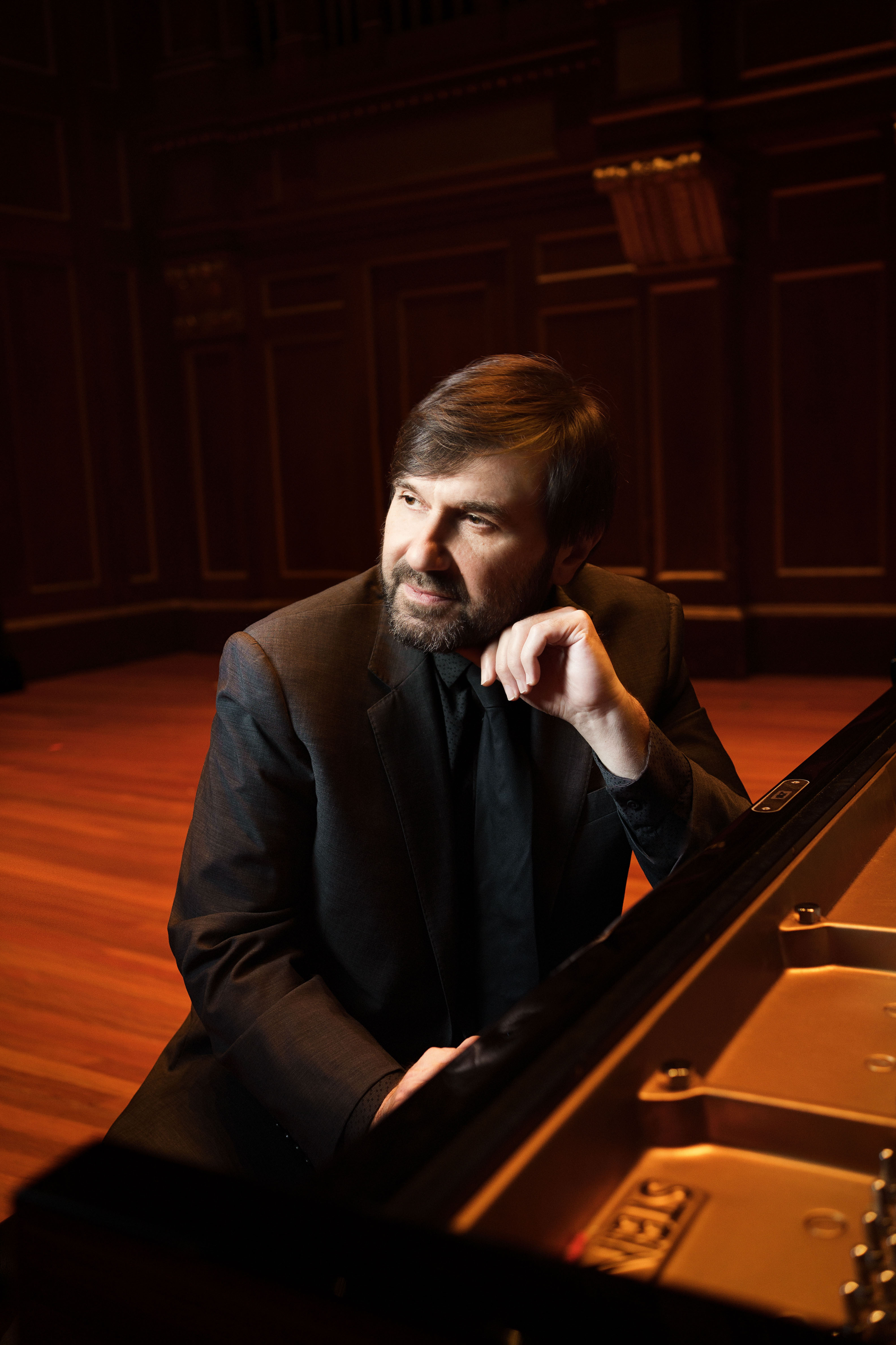 Alexander Korsantia sitting in front of a piano, smiling, with one hand on the keys and one arm resting on the top of the piano