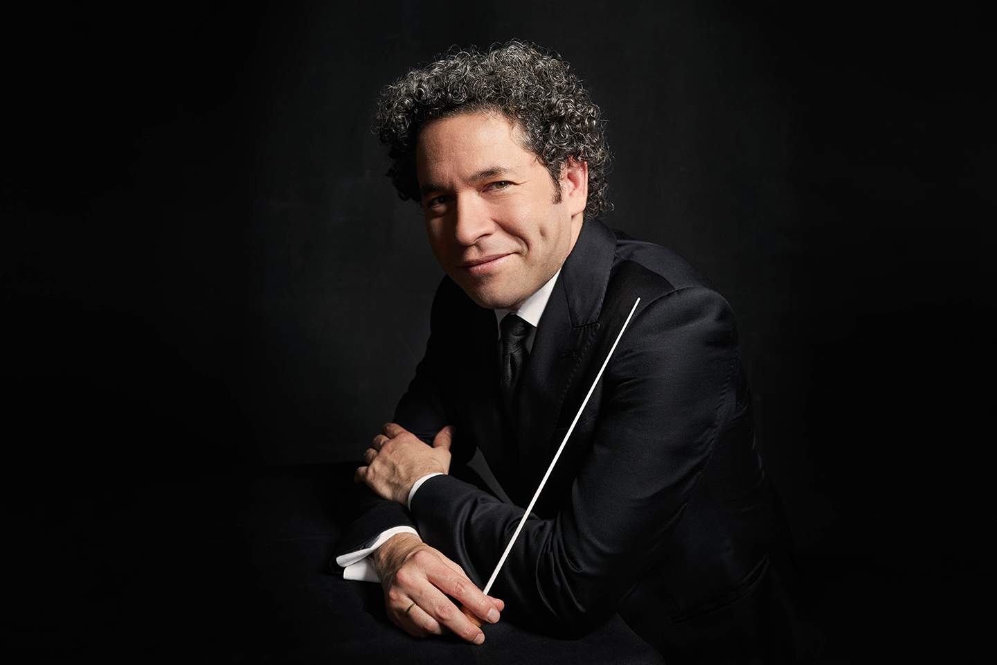 Gustavo Dudamel holding a conductor's baton with his arms crossed in front of him, smiling