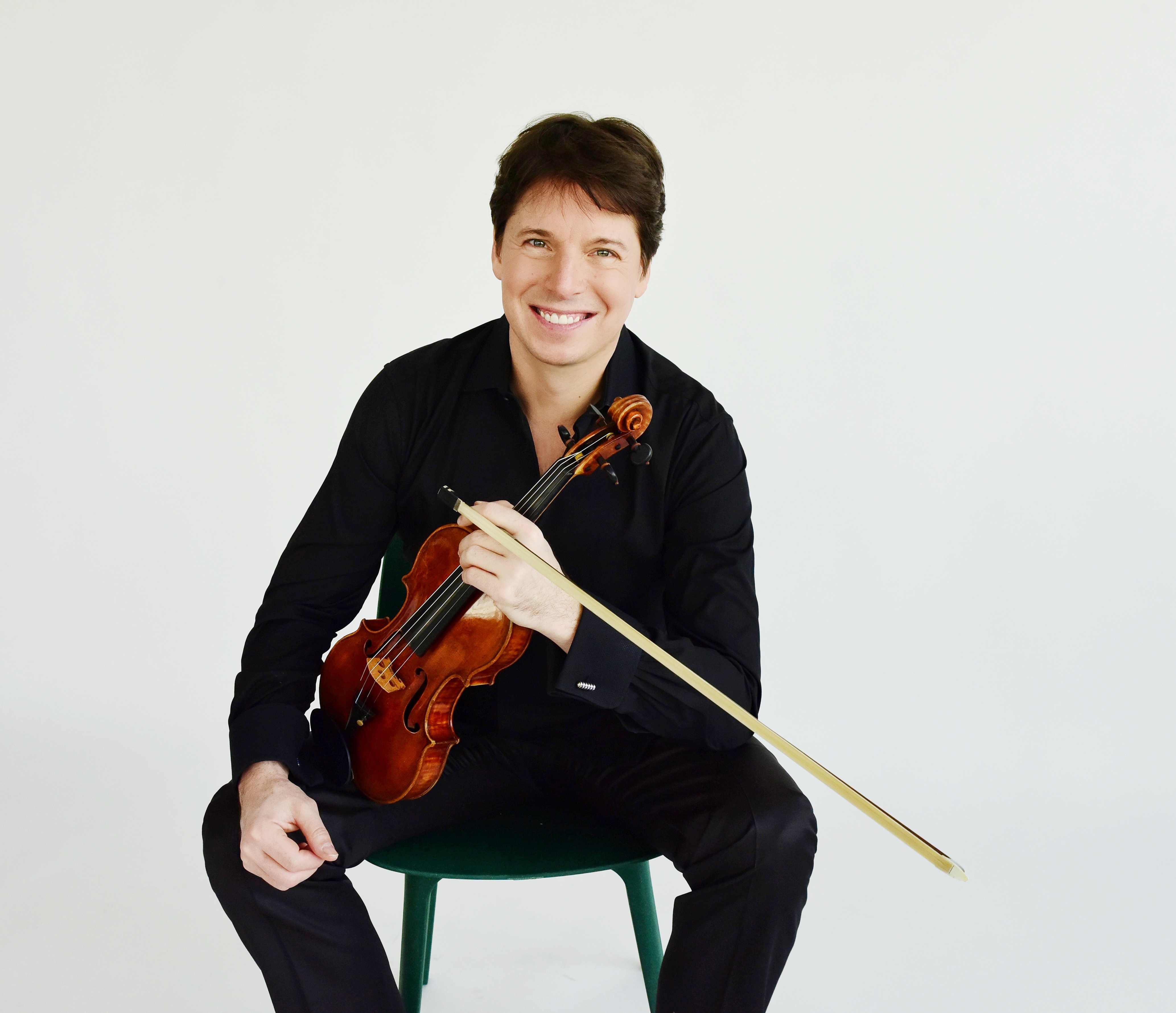 Joshua Bell sitting on a stool, holding his violin and smiling
