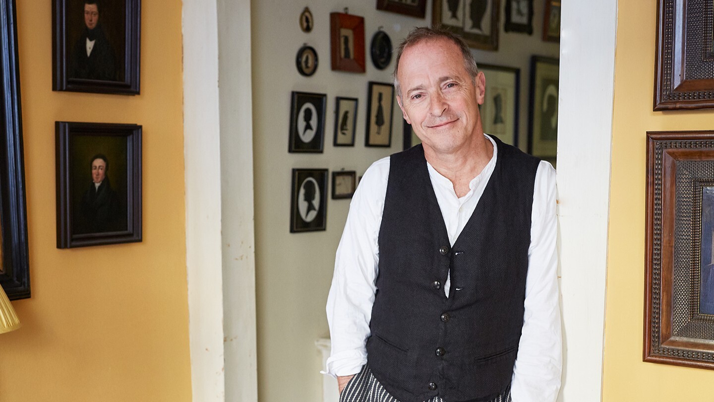 David Sedaris standing in a hallway, surrounded by framed portraits