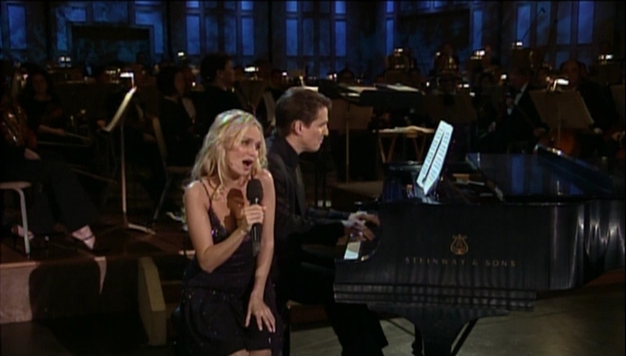 Kristin Chenoweth sings into a microphone while seated on a piano bench; behind her Keith Lockhart plays the piano