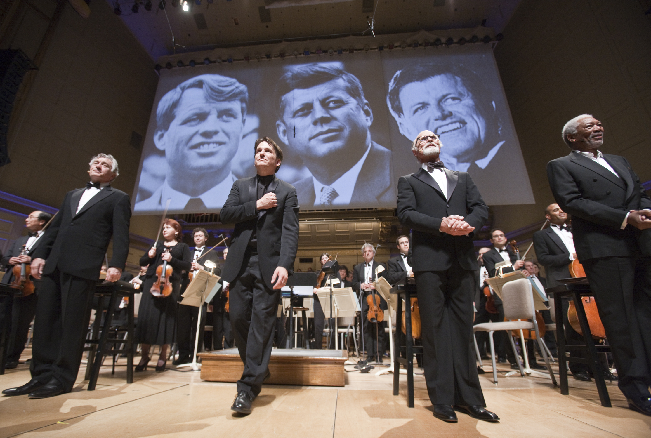 Keith Lockhart stands on the Symphony Hall stage with actors Robert DeNiro, Ed Harris, and Morgan Freeman, with a large image of John, Robert, and Edward Kennedy projected behind them