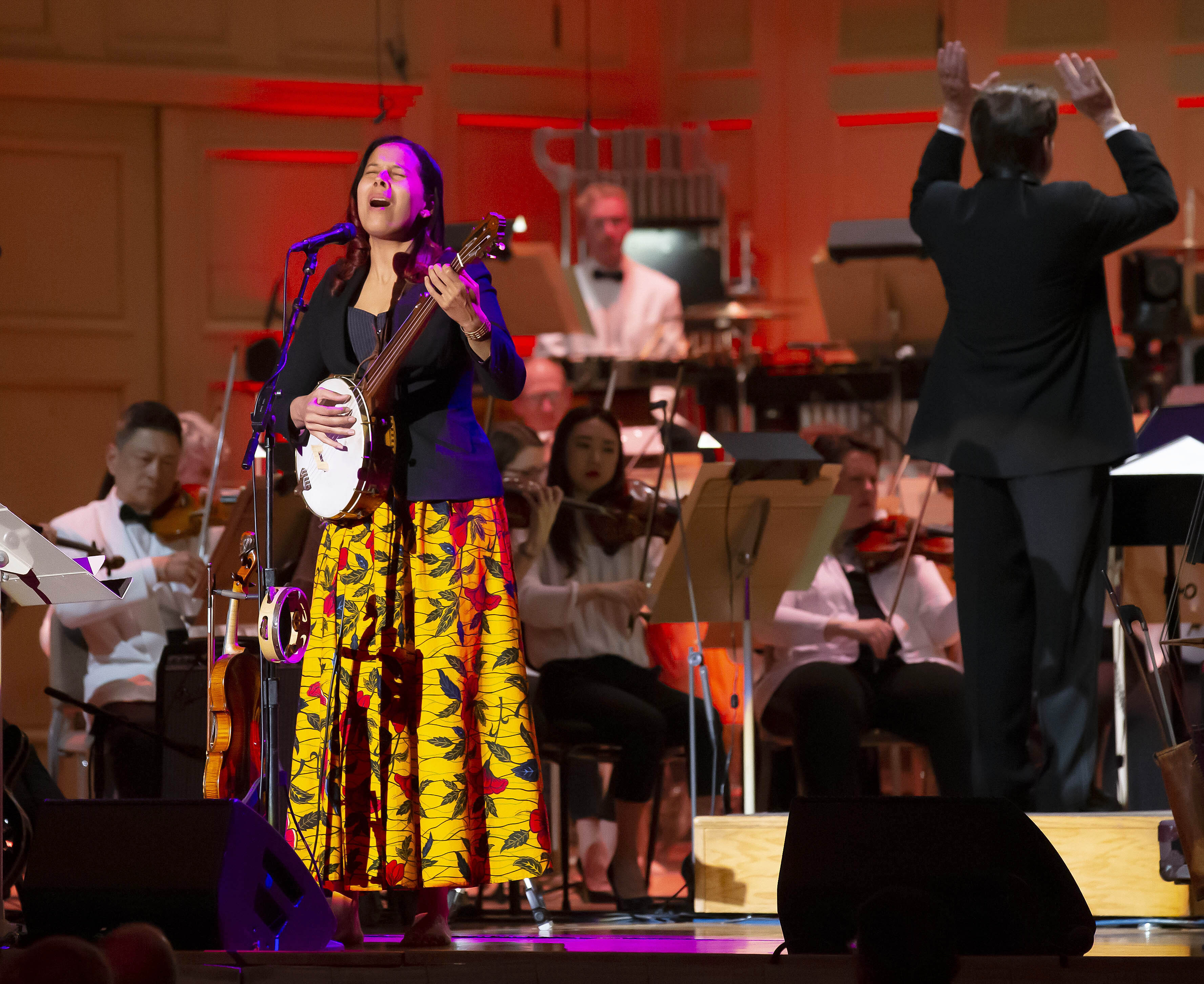 Rhiannon Giddens wears a black jacket and long yellow floral skirt, playing banjo and singing on the Symphony Hall stage as Keith Lockhart conducts the Pops behind her