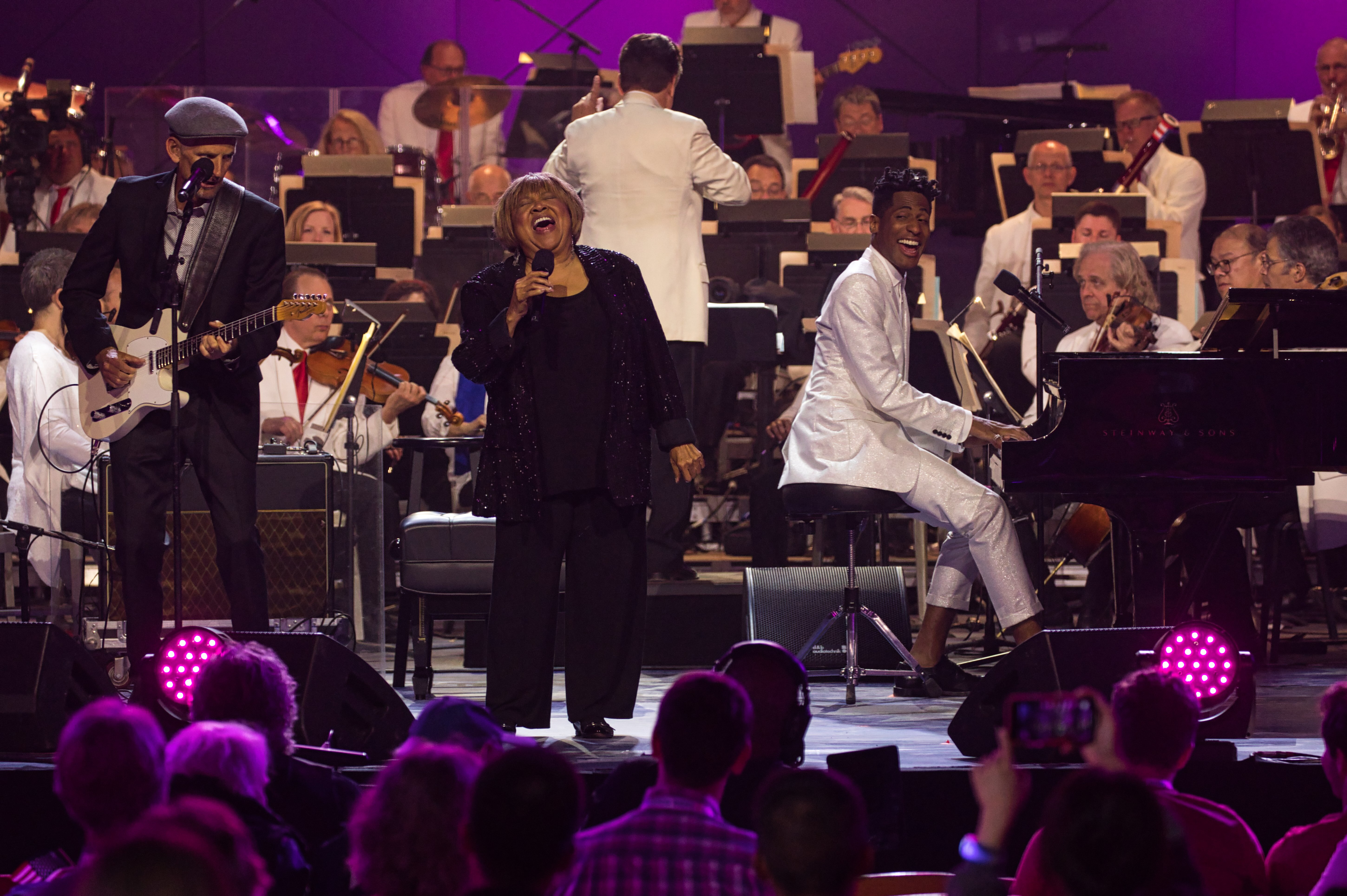 Mavis Staples, wearing all black, sings into a microphone on stage and Jon Batiste, wearing a white suit, plays piano and smiles at the audience. A guitar player accompanies them, and Keith conducts the orchestra behind them.