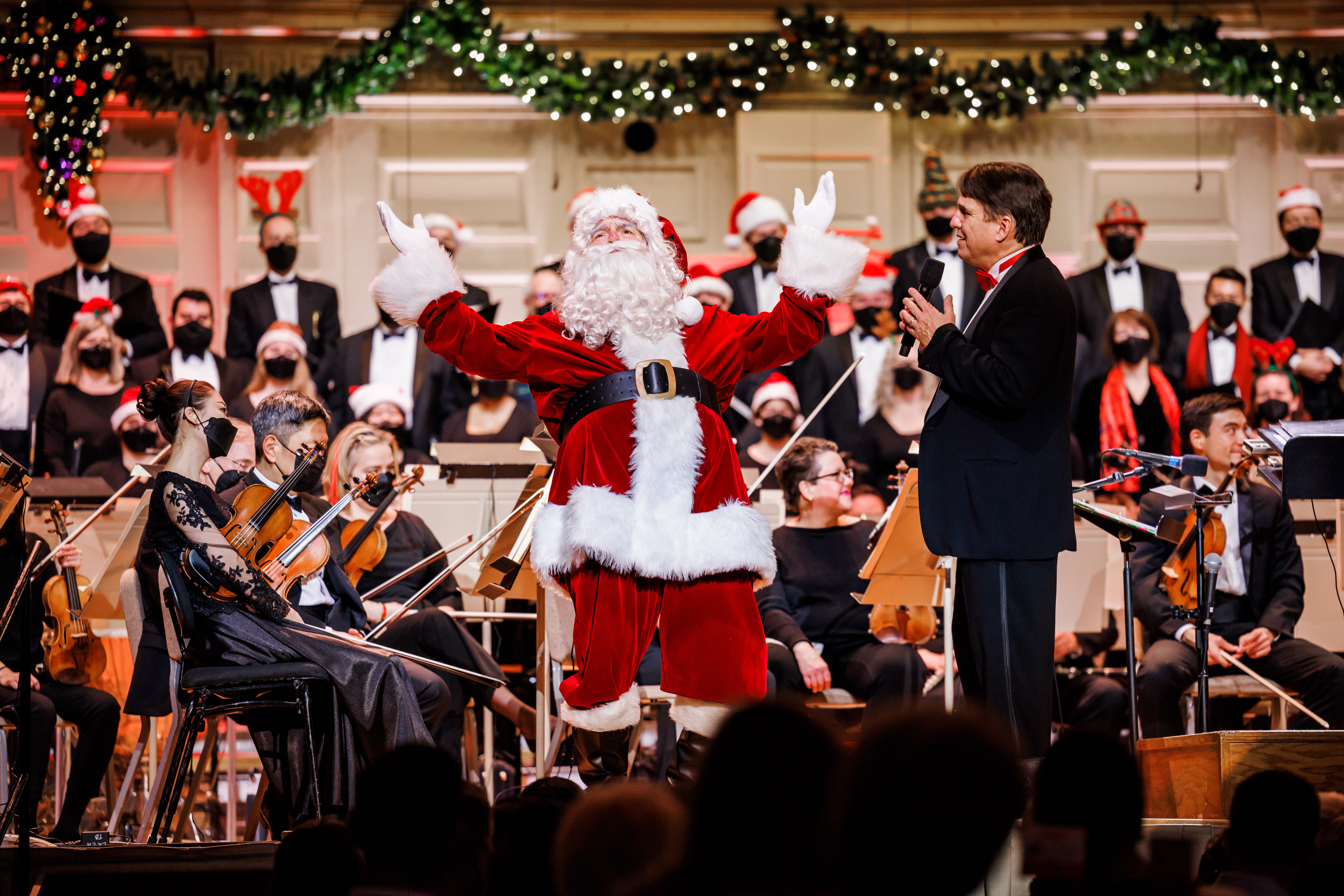Santa Claus stands on the Symphony Hall stage, leans back and raises his arms as Keith Lockhart talks to him