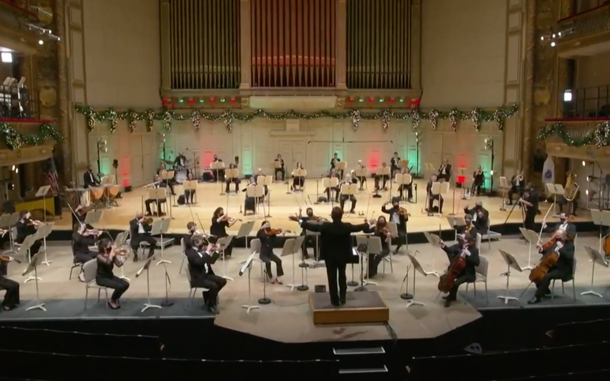The Pops plays on the Symphony Hall stage, wearing all black and sitting far apart to socially distance. The hall is lit with red and green lights and garlands decorate the stage.