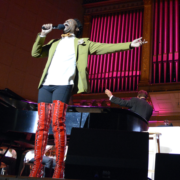 Billy Porter outstretches his arm and sings into a microphone on the Symphony Hall stage, wearing a green suit jacket and tall, red leather boots