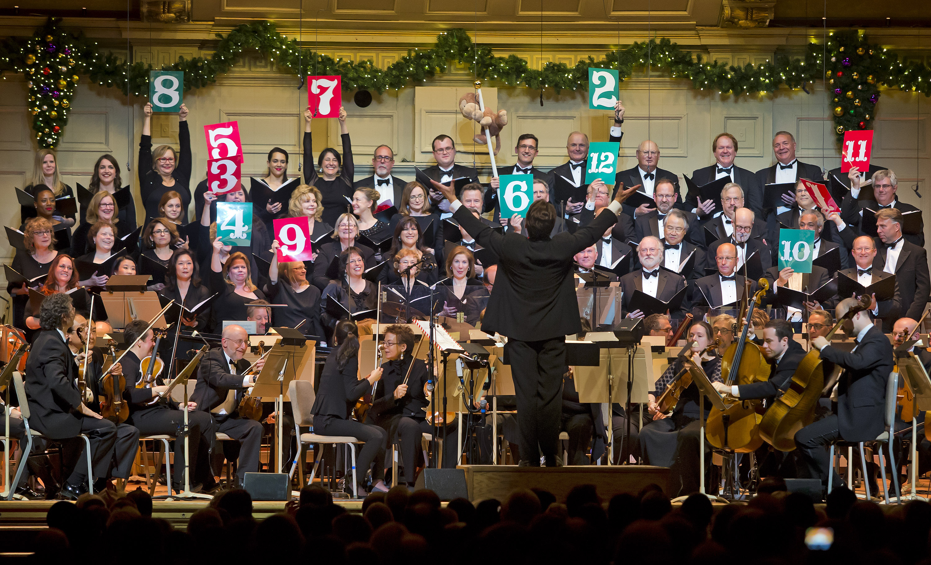 Keith Lockhart stands on the Symphony Hall podium with arms outstretched in front of the Tanglewood Festival Chorus, the singers smiling as some hold red and green signs of numbers