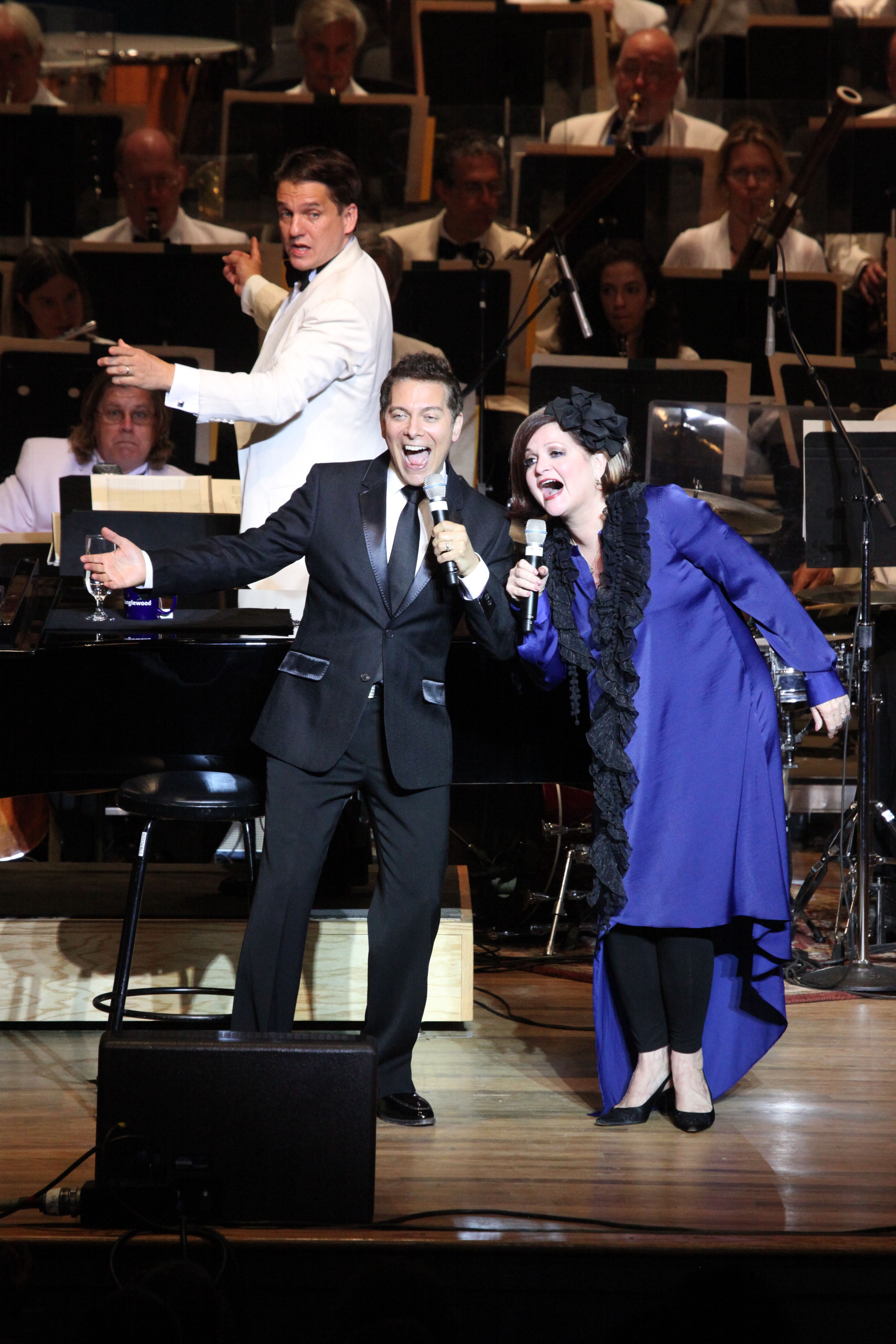 Michael Feinstein wears a black suit and Faith Prince wears a long blue dress with a black scarf. They smile and sing together on stage, with Keith Lockhart conducting behind them