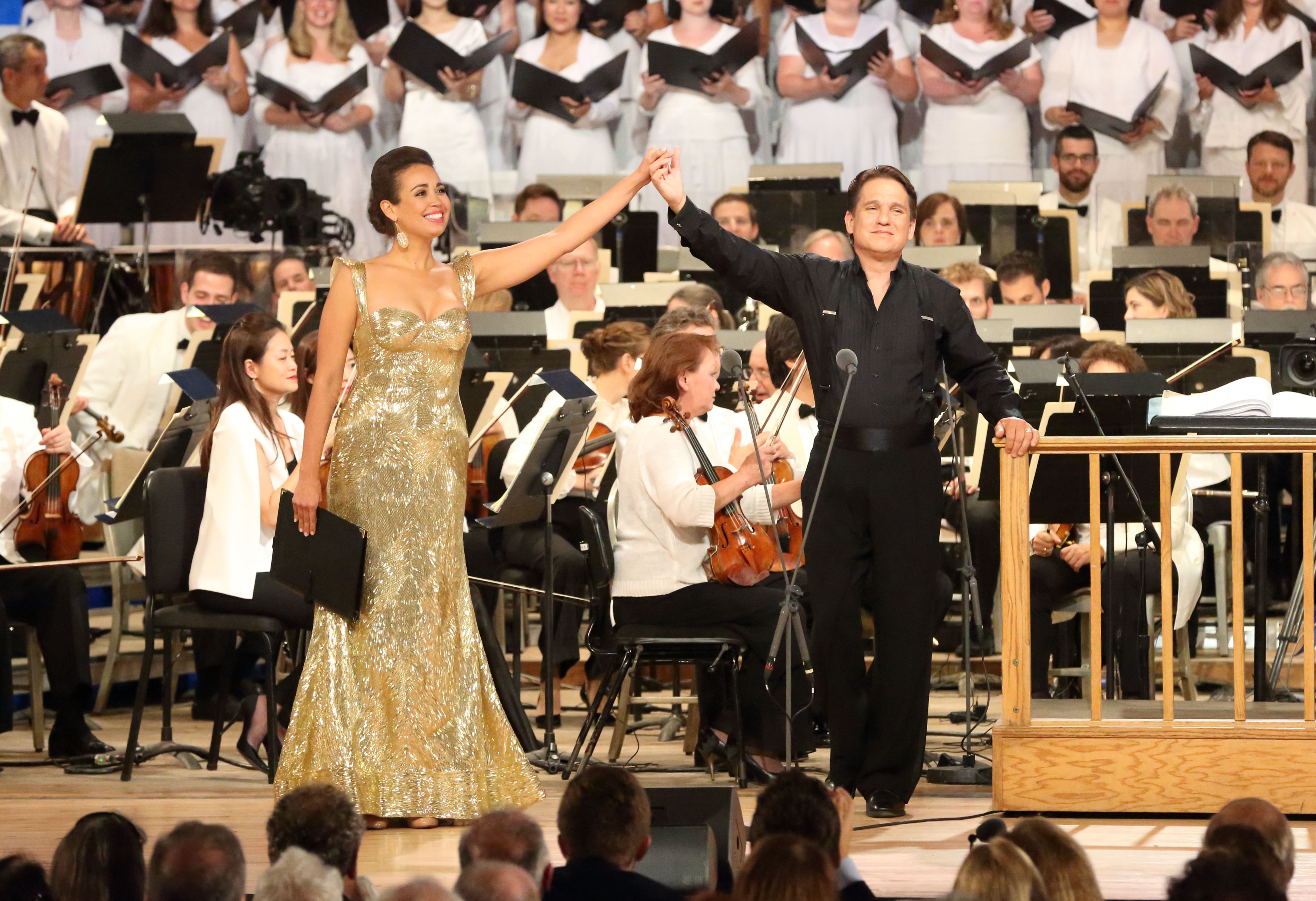 Nadine Sierra wears a sparkly, gold gown, and Keith Lockhart wears a black dress shirt and pants as the two hold hands on stage, lifting them high before taking a bow
