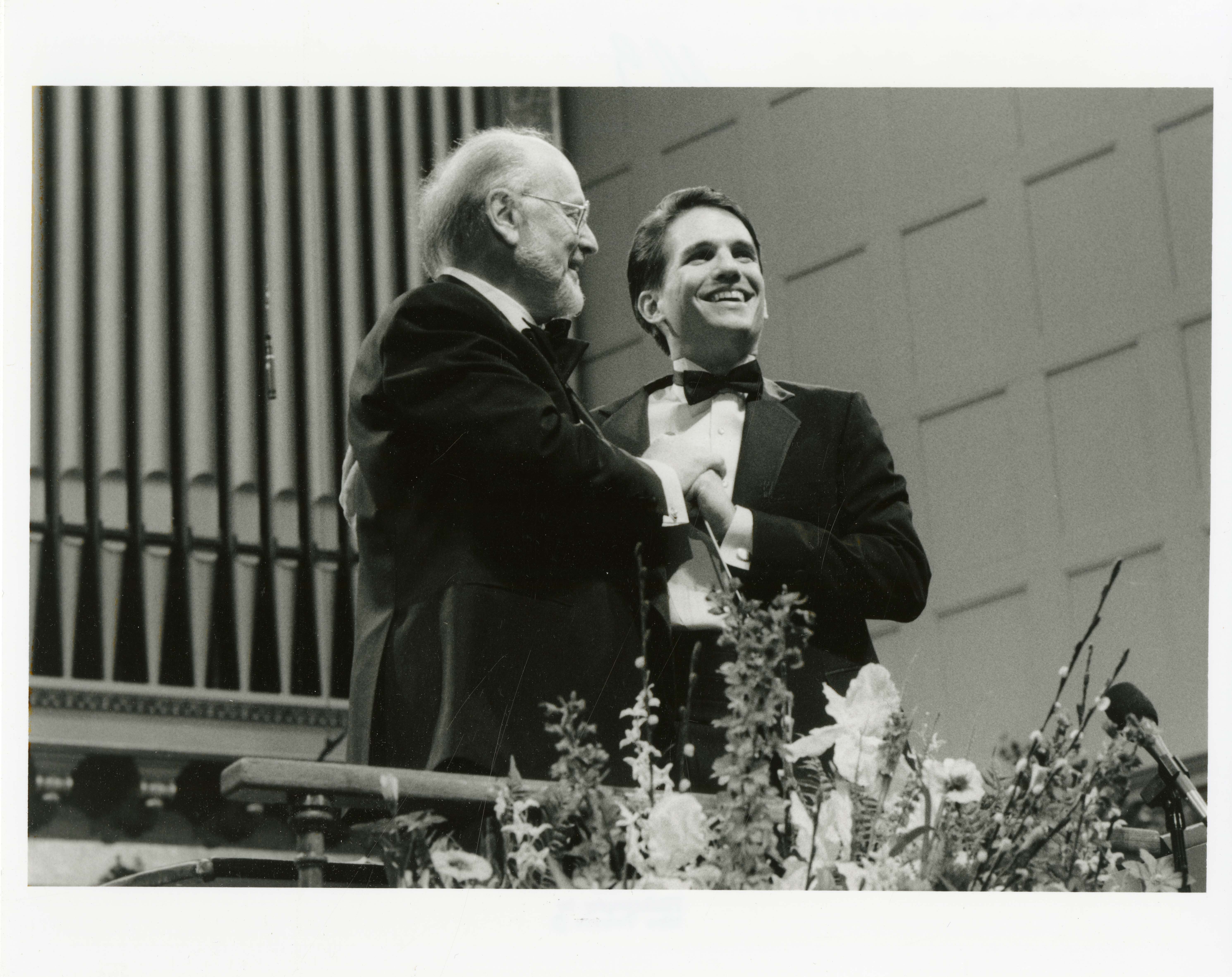 Black and white photo of John Williams passing the baton to Keith Lockhart on the Symphony Hall stage.