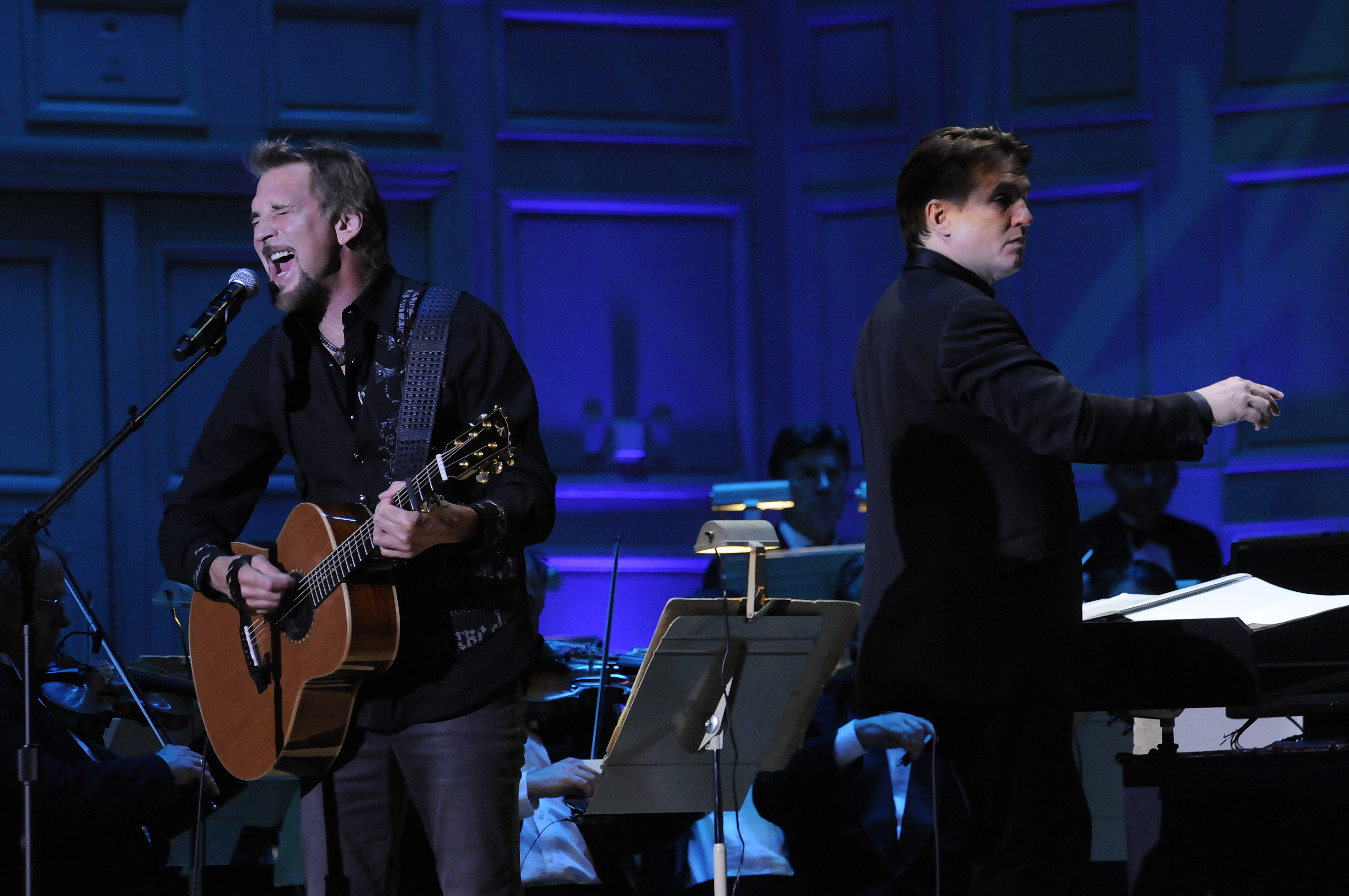Dressed in dark jeans and a black shirt, Kenny Loggins plays acoustic guitar and sings on the Symphony Hall stage, with Keith Lockhart conducting the Pops behind him