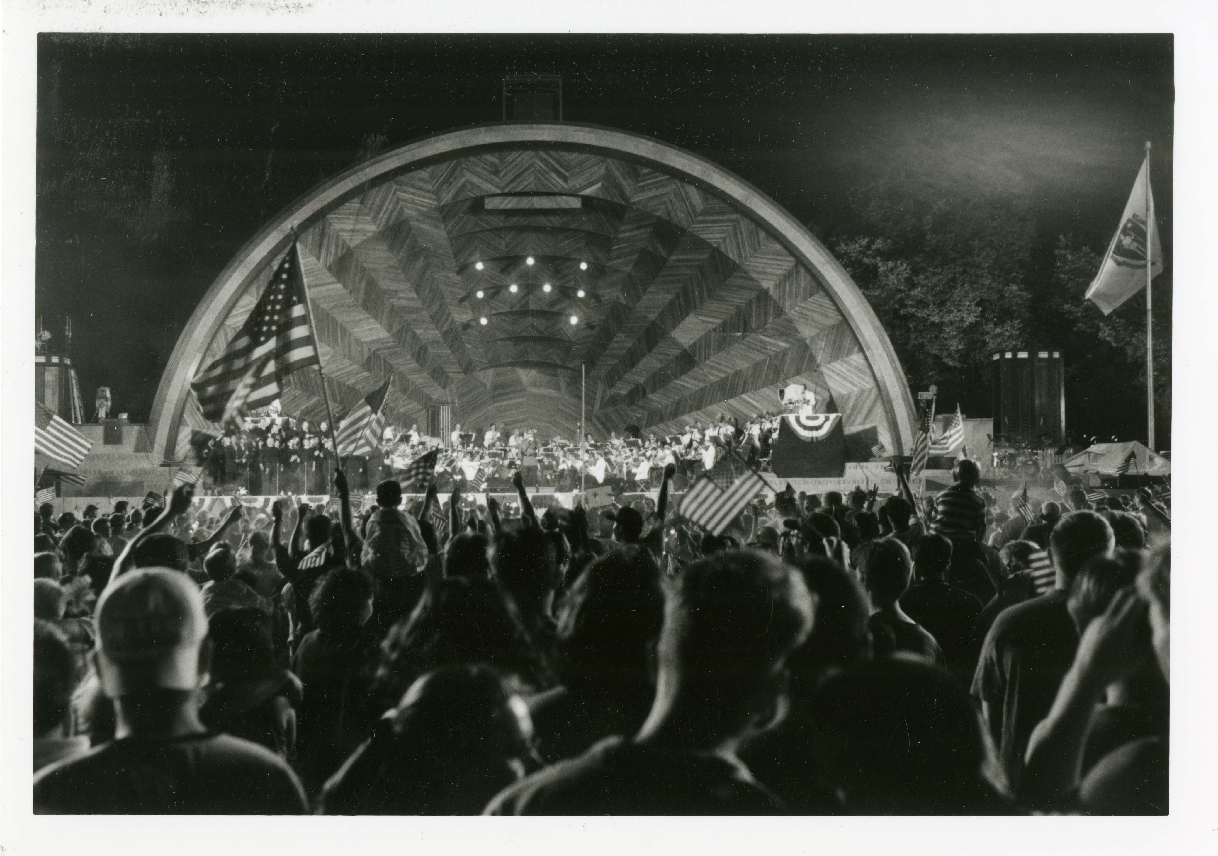 A black-and-white photo of a large crowd waving American flags in front of the Hatch Shell, a large stage with a semicircular arch
