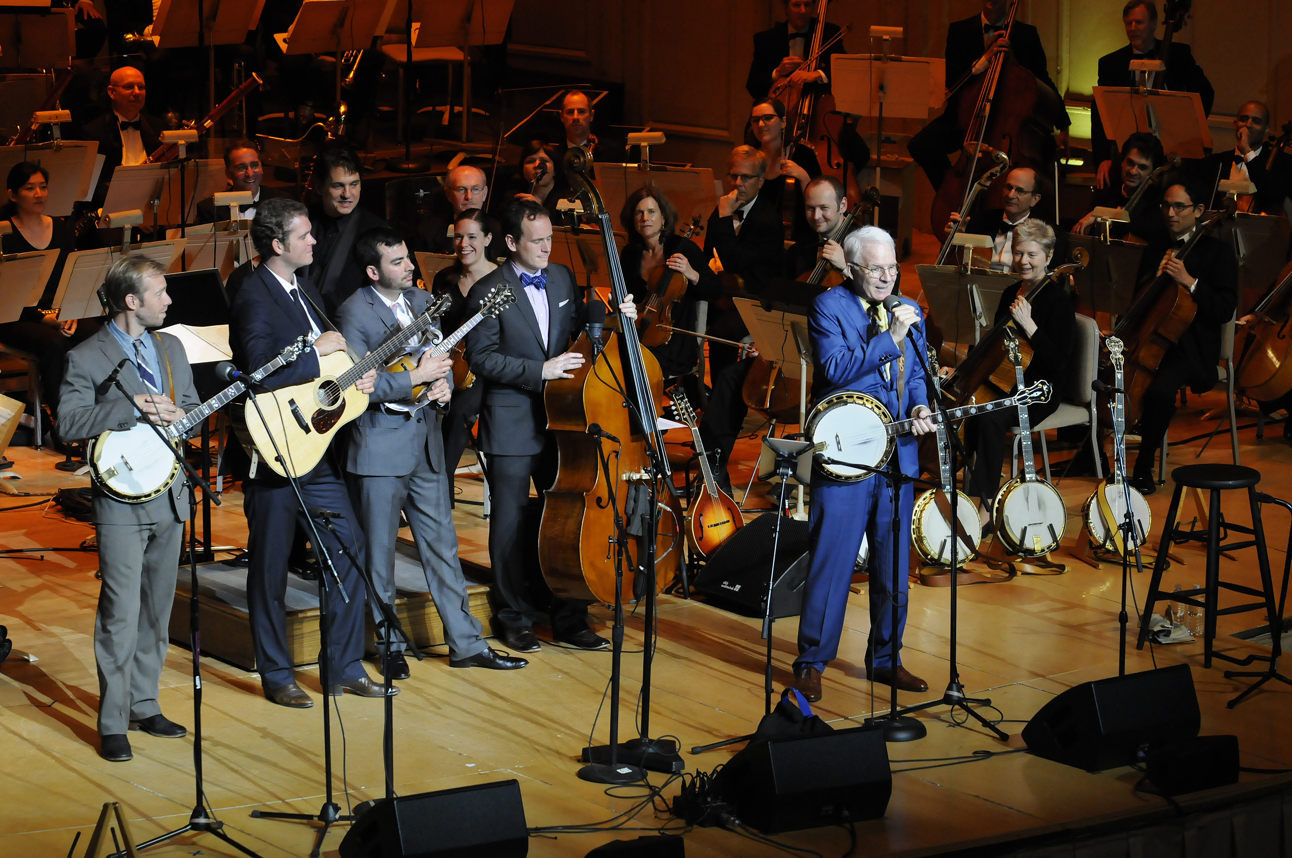 Dressed in a blue suit, Steve Martin holds a banjo and speaks into a microphone from the Symphony Hall stage, as the musicians behind him watch