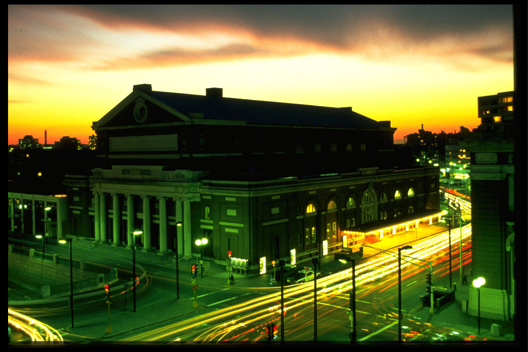 Exterior of Symphony Hall as the sunsets in the background, with blurred traffic and car lights