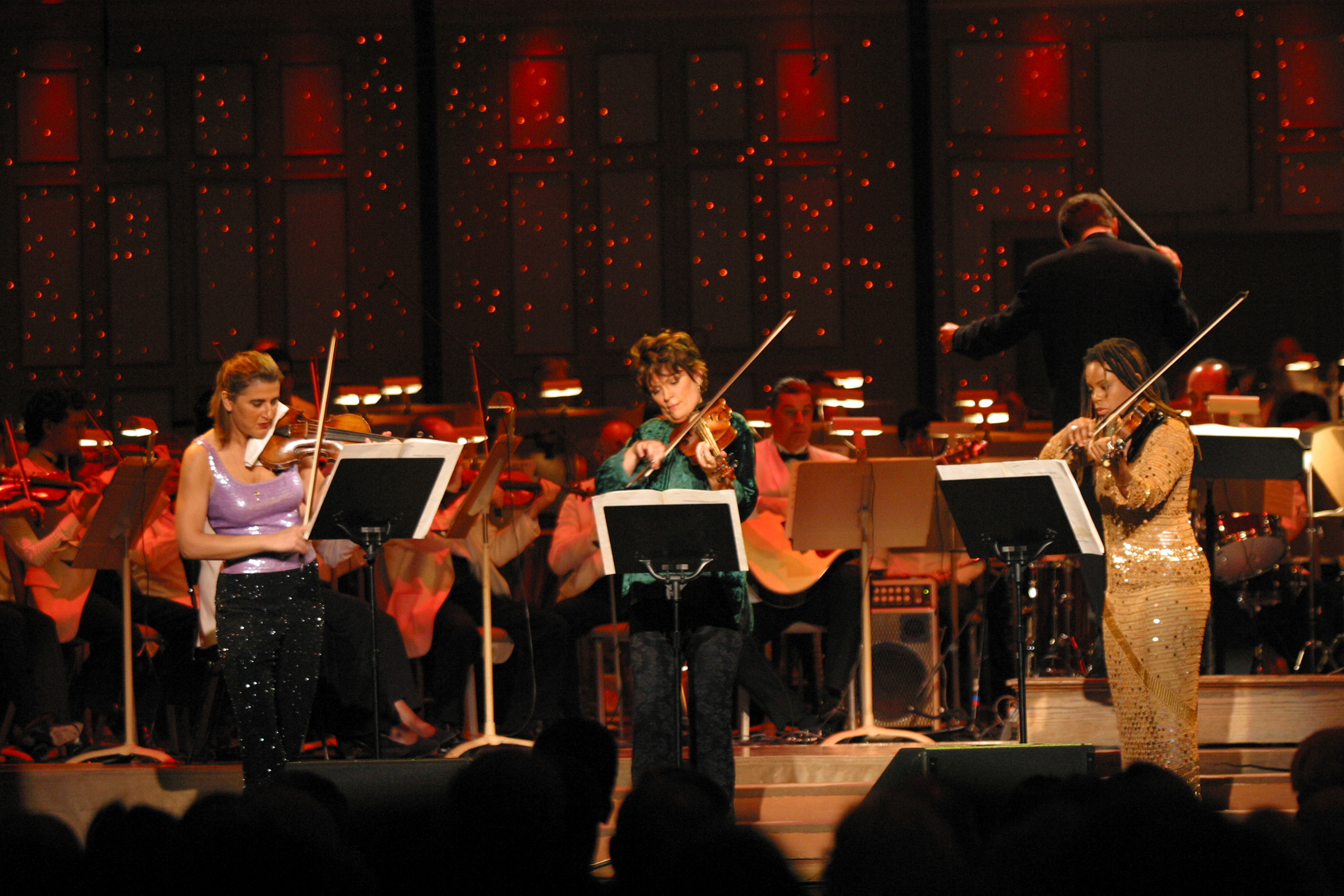 Three violinists stand on stage in front of Keith Lockhart conducting in the background