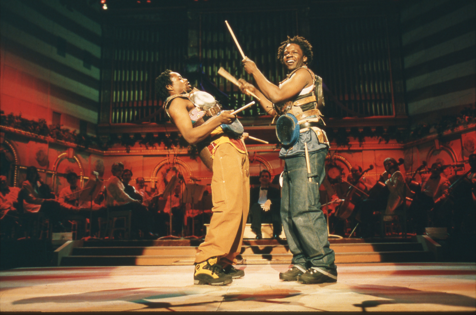 One musician with percussion instruments strapped to his torso faces the audience and raises two drum sticks while another musician bends away from him